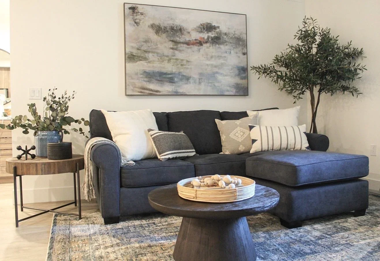 Living room with a dark blue sectional sofa adorned with white and patterned throw pillows, a wooden coffee table with a decorative tray, a round side table with a vase of greenery and cups, a large abstract painting on the wall, and a tall potted pl