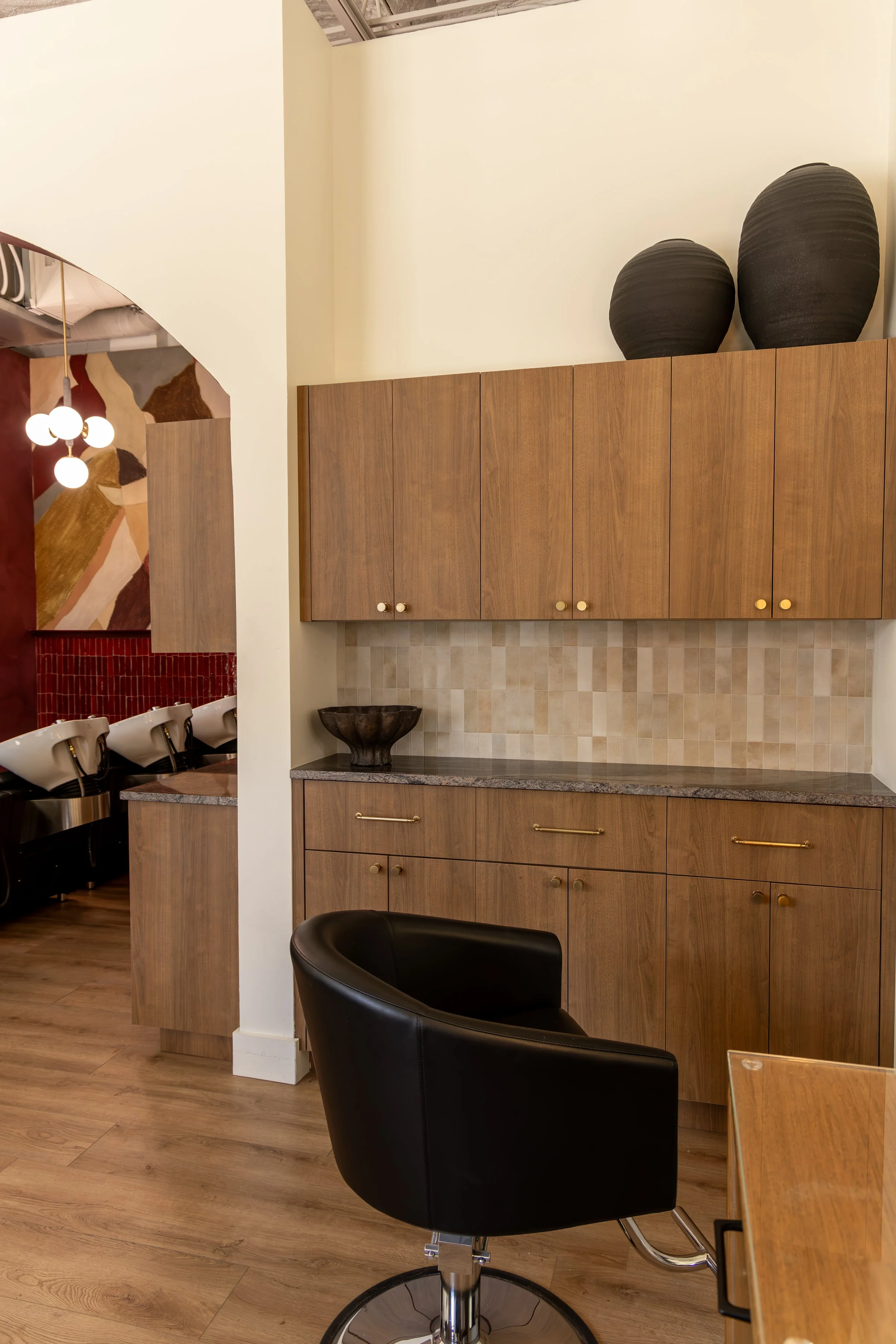 Interior of a modern coffee shop or cafe with wooden cabinets, black decorative vases on top, and a black leather swivel chair in the foreground. The space has warm lighting and a clean, minimalist design.
