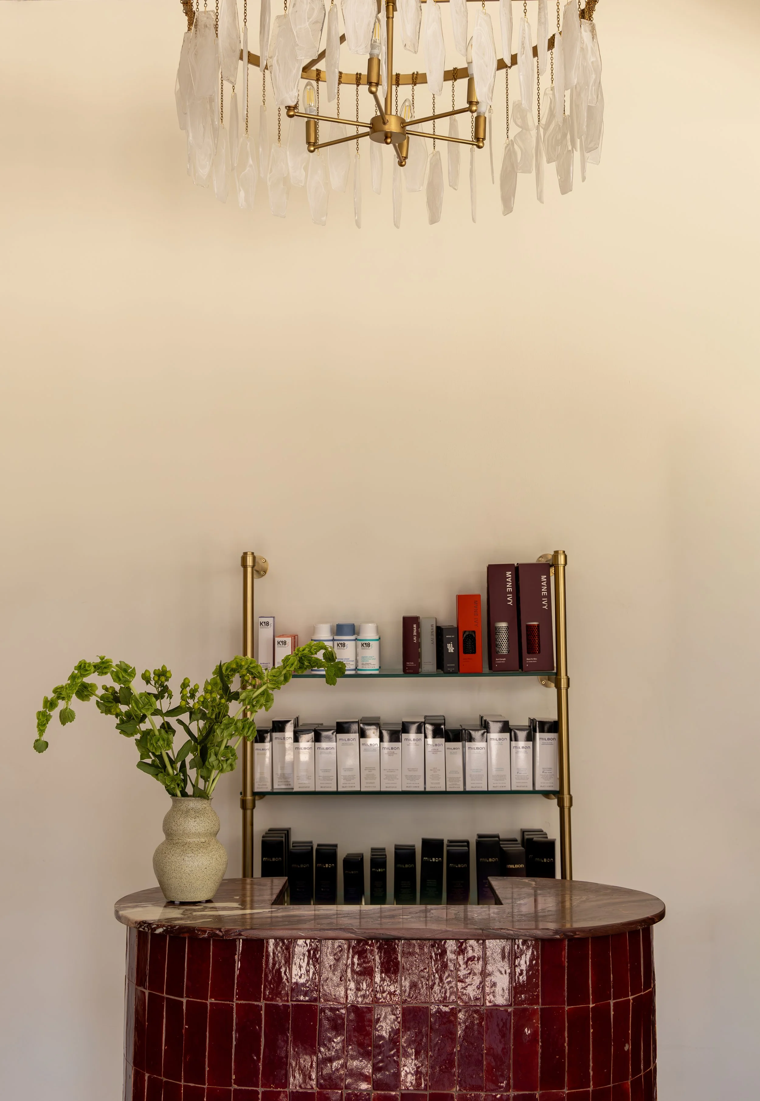 A reception desk with a round, dark red tiled front and a marble top, with a large green potted plant on the left. Behind it, a gold-framed wall shelf system displays various boxed products. Above, a decorative chandelier with clear glass pieces hangs from the ceiling.