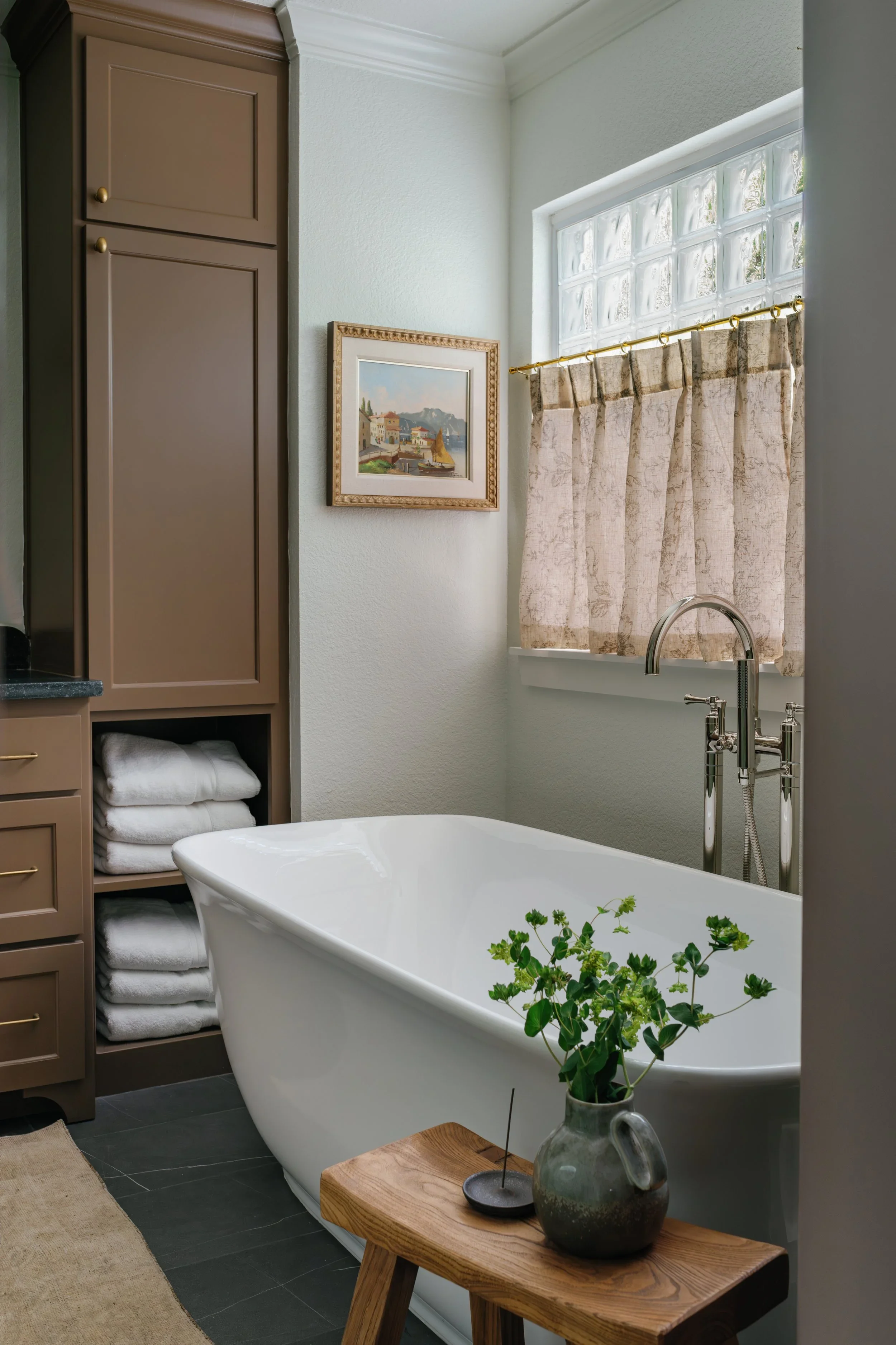 A bathroom with a white bathtub, a wooden side table with a plant in a vase, a wall with beige cabinets, a framed painting, a window with frosted glass blocks and beige curtains, and a black tiled floor.