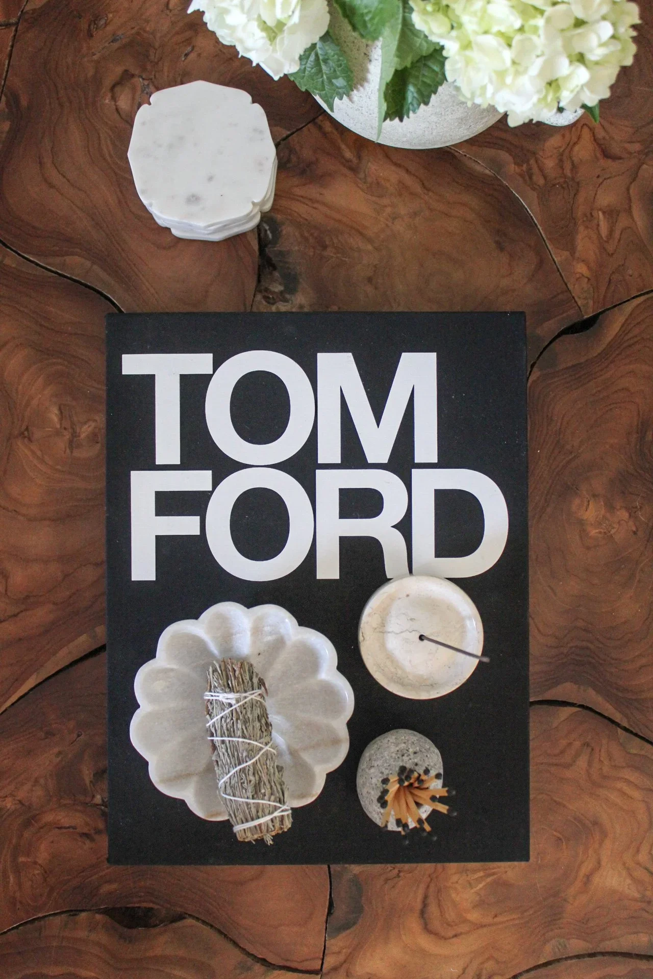 A black book with the words "TOM FORD" in white, surrounded by candles, dried lavender, and decorative items on a wooden table with a bouquet of white hydrangeas in a gray pot.