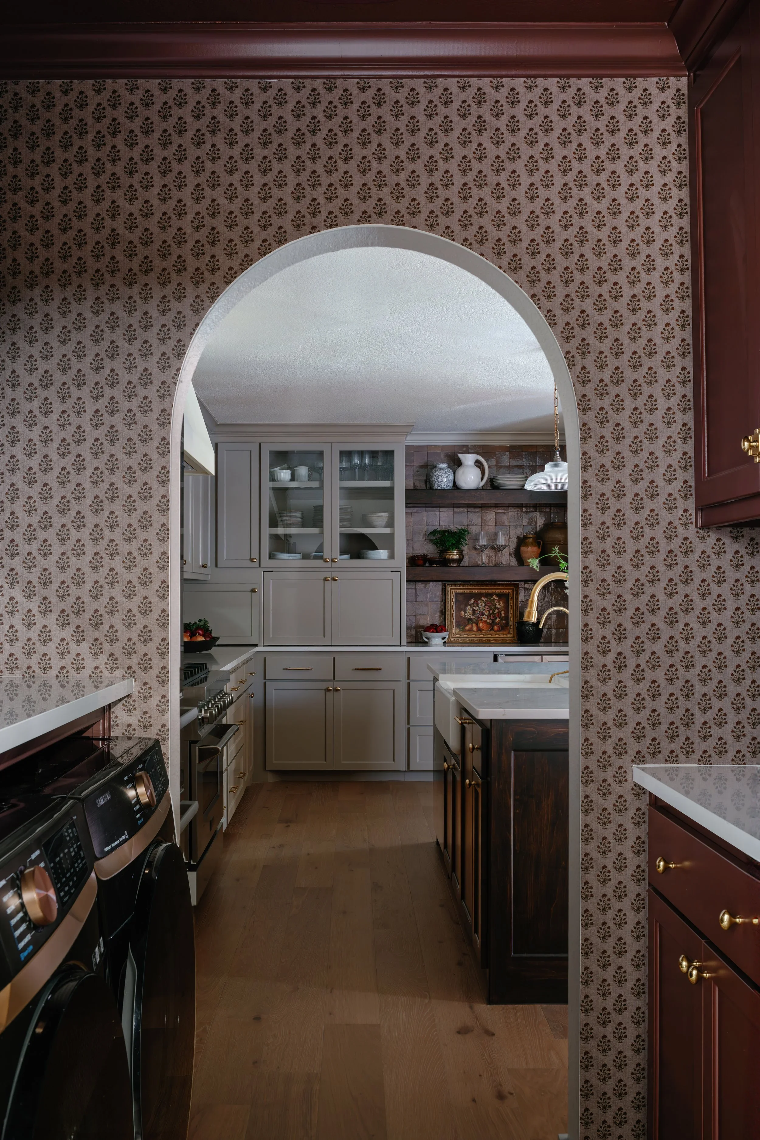 A view through an arched doorway into a kitchen with a patterned wallpaper on the wall, a white cabinet with glass doors, and open wooden shelves with decorative items including white vases, plates, and framed artwork. The kitchen has a white farmhou