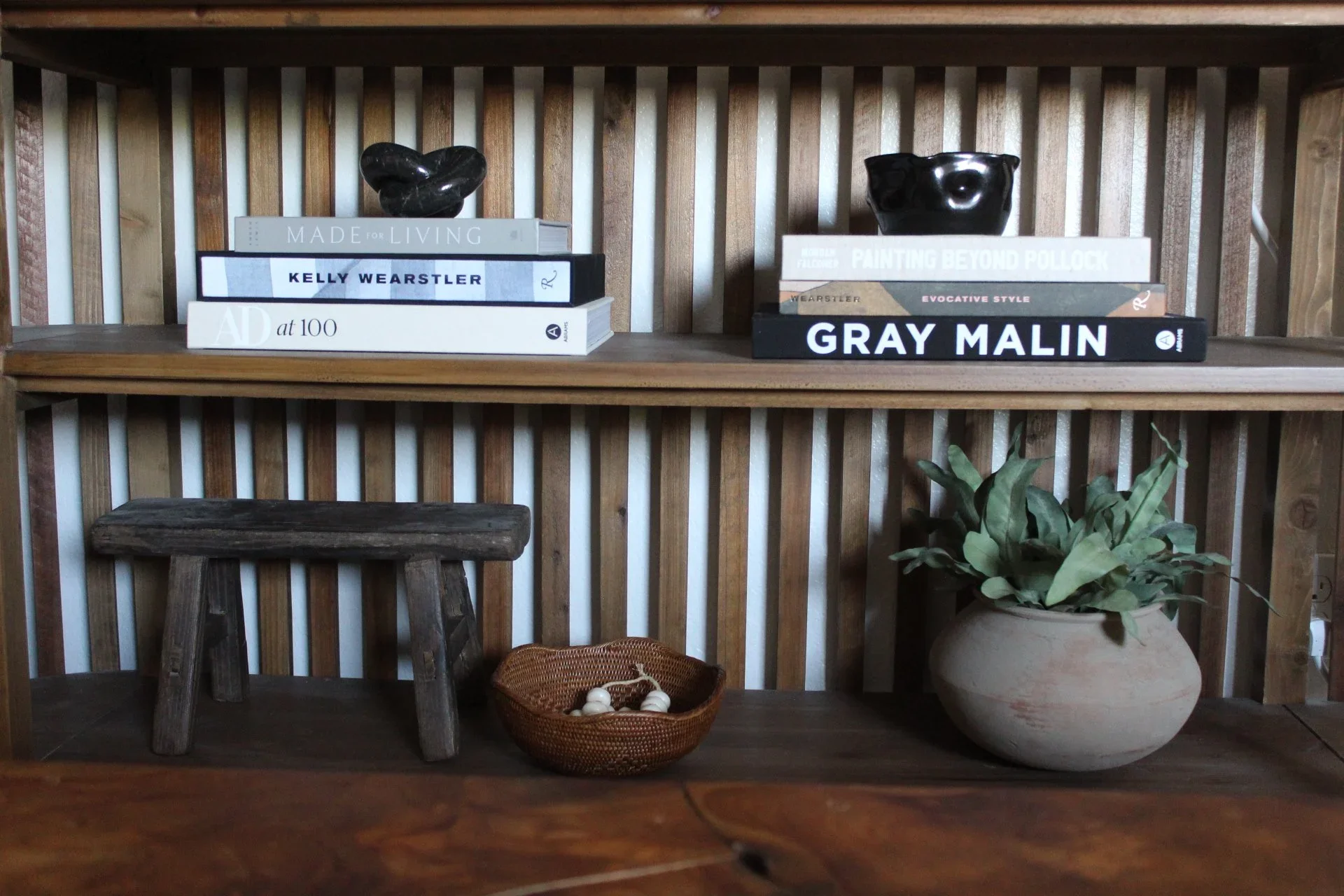 A wooden shelf with two stacks of books and two black decorative bowls on top. Below, there is a wooden stool and a large pot with green plants. The background features a wooden slat wall.