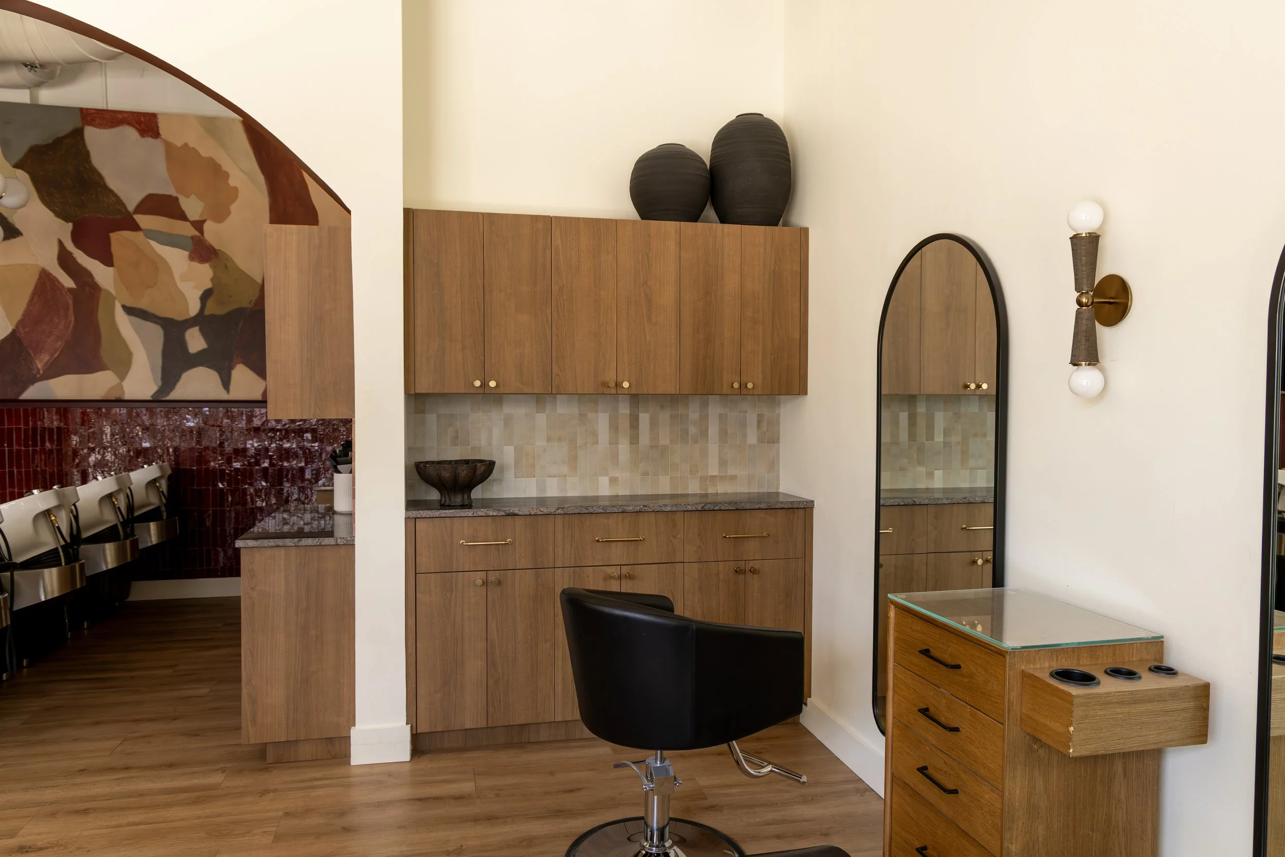 Interior view of a room with wooden cabinets, a black chair, a glass-top wooden cabinet, and decorative black vases on top of the cabinets. There are mirrors on the wall and a wall sconce with dual white light bulbs. Part of the adjacent room with chairs and a mosaic wall is visible.