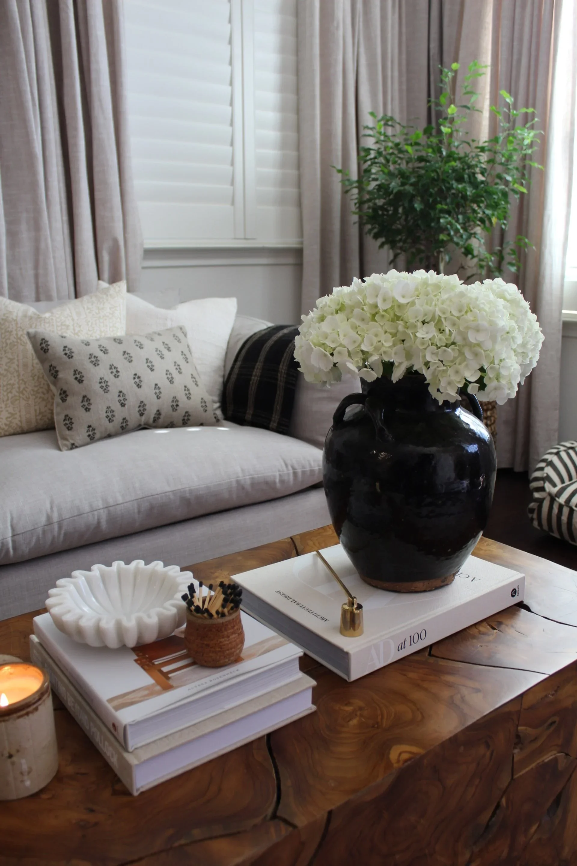 A living room with a gray couch and decorative pillows, a black vase with white hydrangea flowers on a wooden coffee table, surrounded by stacked books, candles, and small decor items, with curtains and a potted plant in the background.