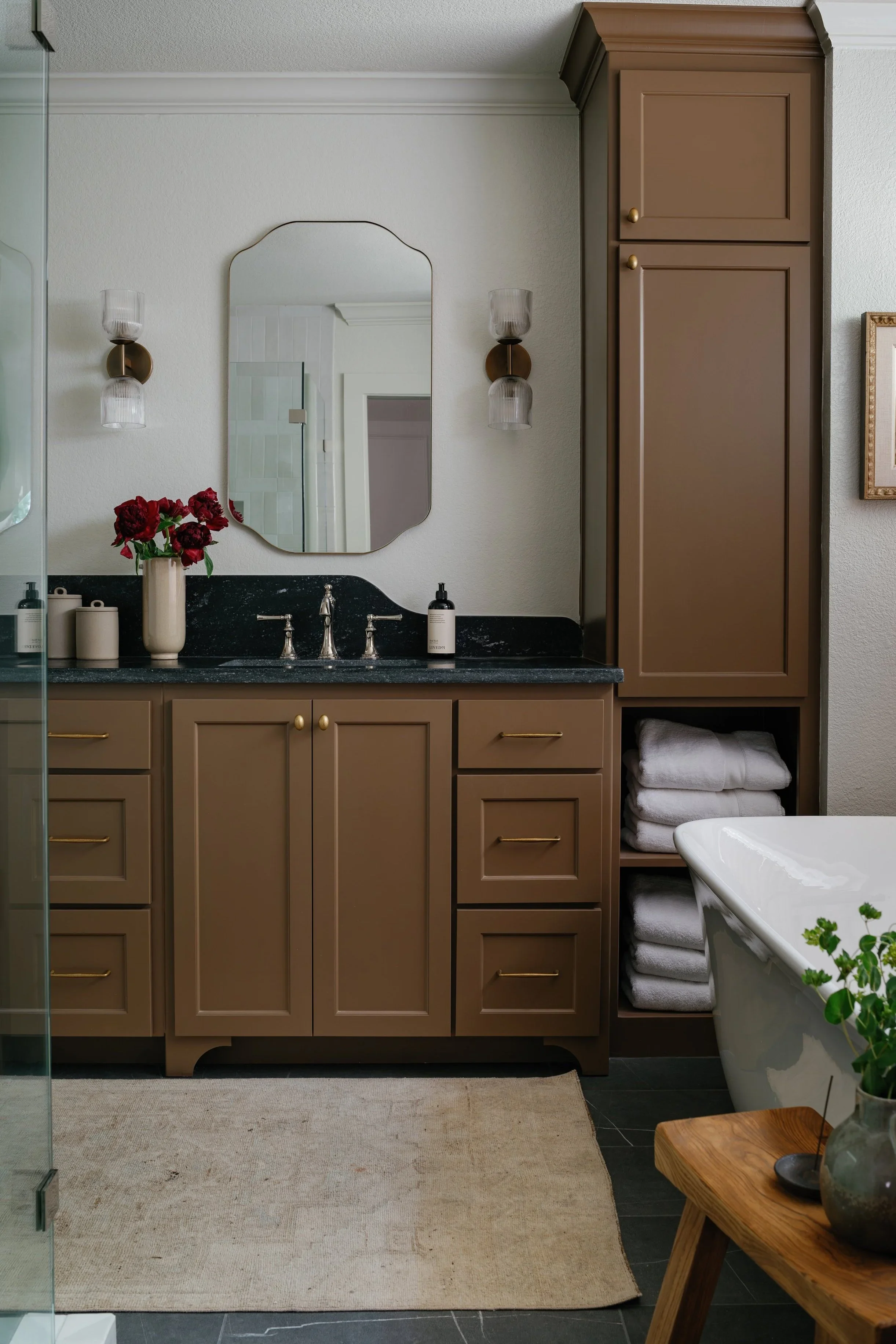 A bathroom with beige cabinets, a black countertop, a mirror, and wall-mounted light fixtures. There are towels stored in an open cabinet and a bathtub with a plant on nearby table.