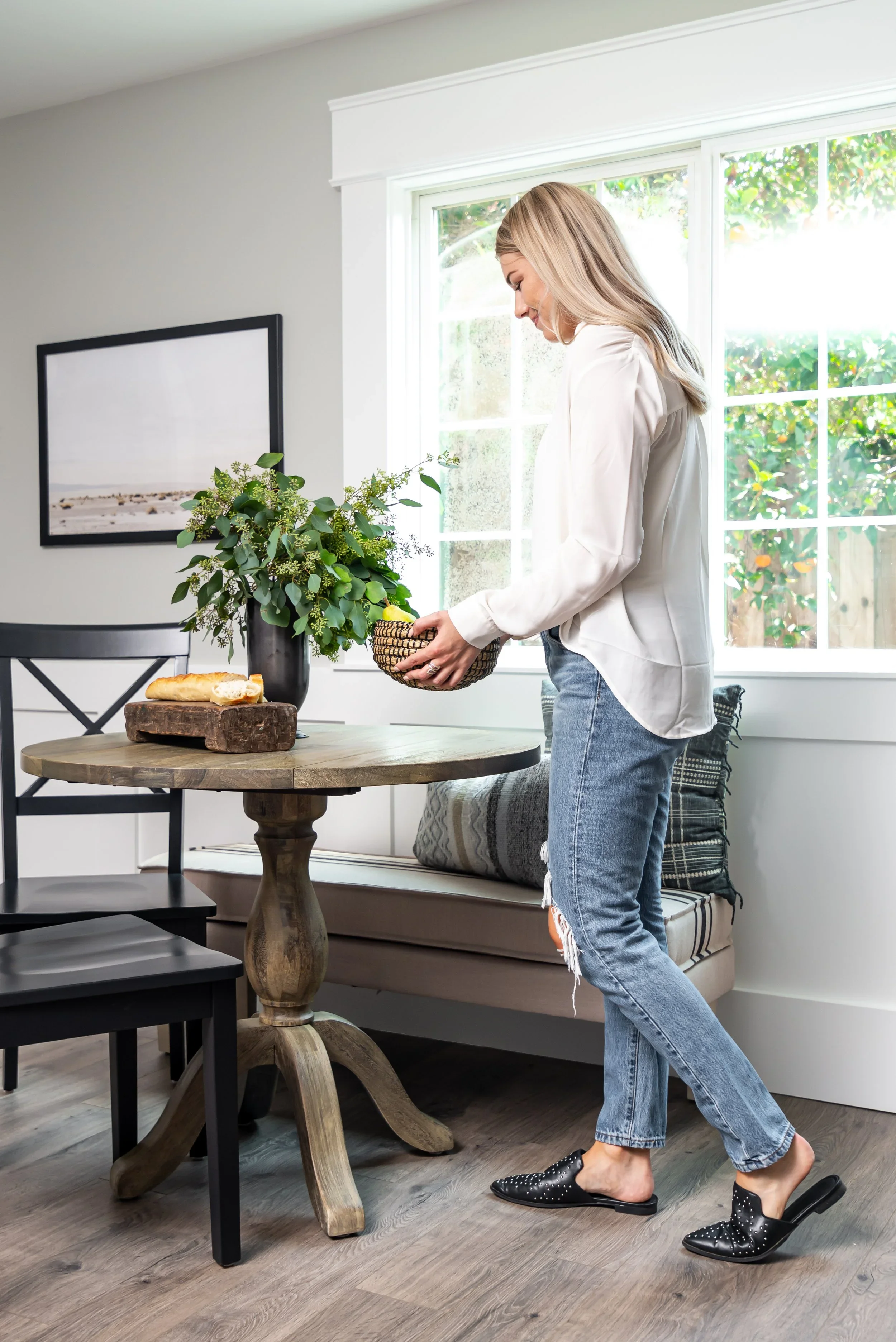 A woman is standing in a bright dining room, holding a basket, wearing a white jacket, jeans with a ripped knee, and black slip-on shoes with white polka dots. There is a wooden table with a loaf of bread and a vase of greenery, and a beige bench wit