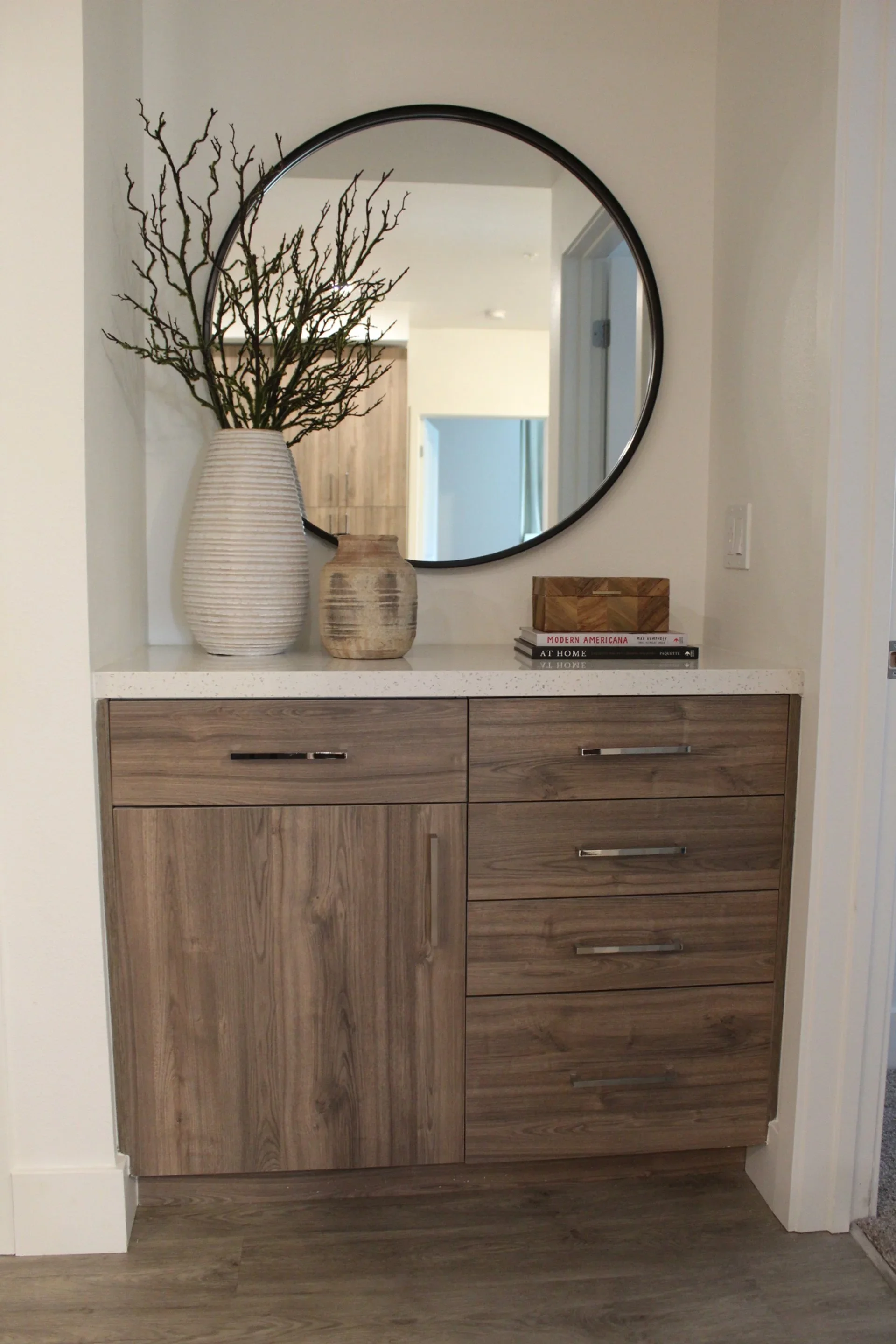 A wooden cabinet with five drawers and one door, topped with a white counter. Above the cabinet, a round mirror is mounted on the wall. On the counter, there are two decorative vases, one large with bare branches and one smaller, along with a stack o