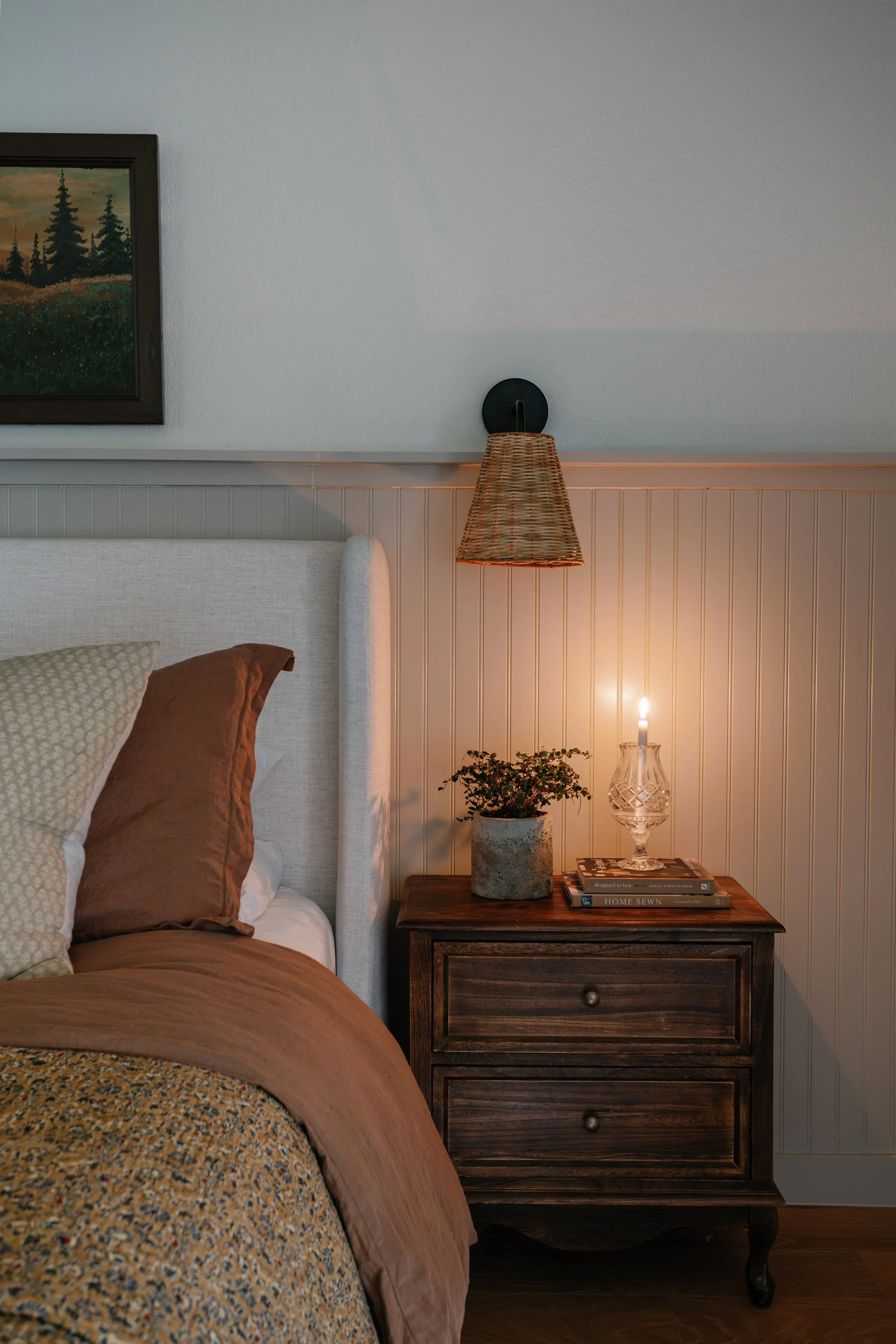 A bedroom corner with a bed featuring brown and patterned pillows, a wooden nightstand with books, a lamp, and a potted plant. Above the bed is a wall-mounted lamp with a woven shade, and a framed landscape painting of trees on the wall.