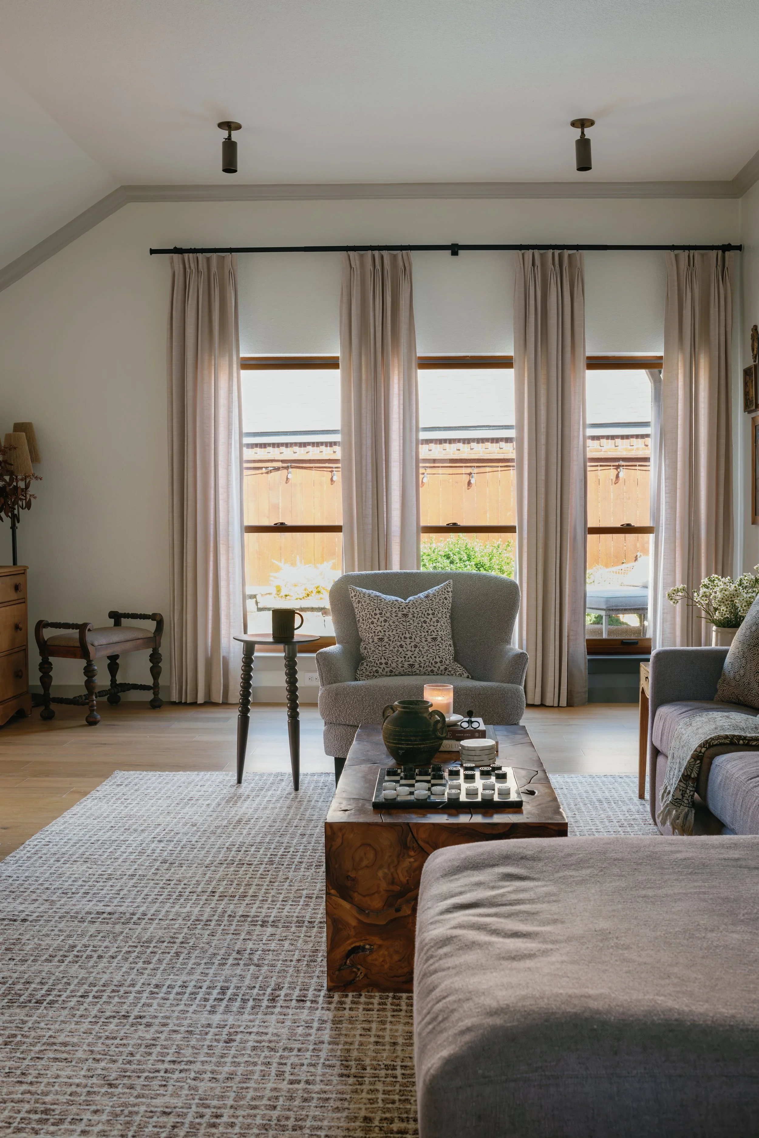 Living room with large windows, beige curtains, a grey armchair with a decorative pillow, a rustic coffee table with a decorative vase and chessboard, a patterned area rug, and neutral-toned decor.