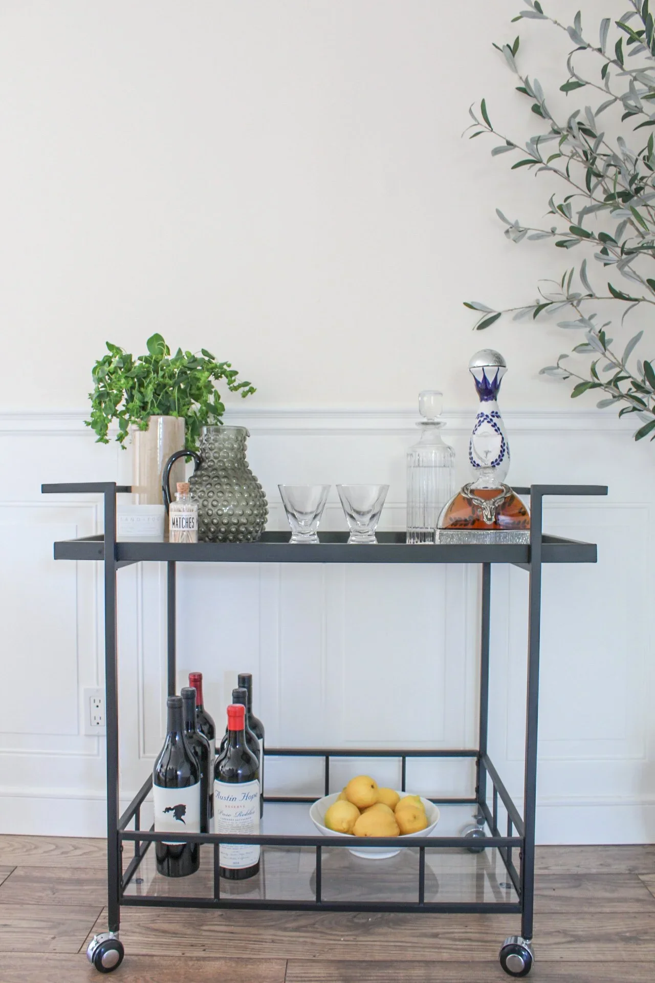 A black metal bar cart with two shelves on wheels, holding wine bottles, a bowl of lemons, and various glassware and decorative items, set against a white wall with wall paneling.