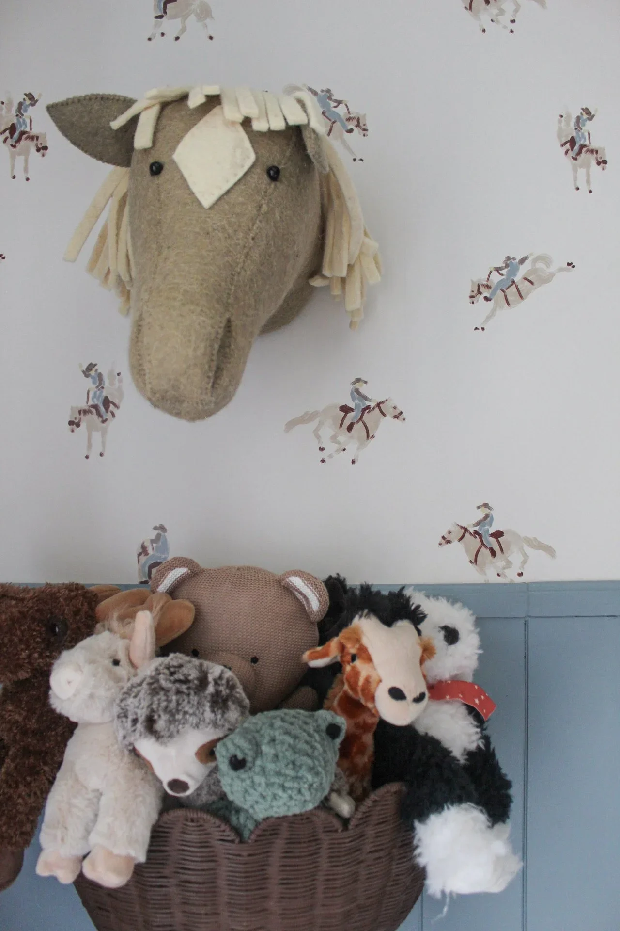 A collection of stuffed animals, including a plush horse hanging on the wall and various plush animals in a basket below, with a wallpaper featuring cowboys riding horses in the background.