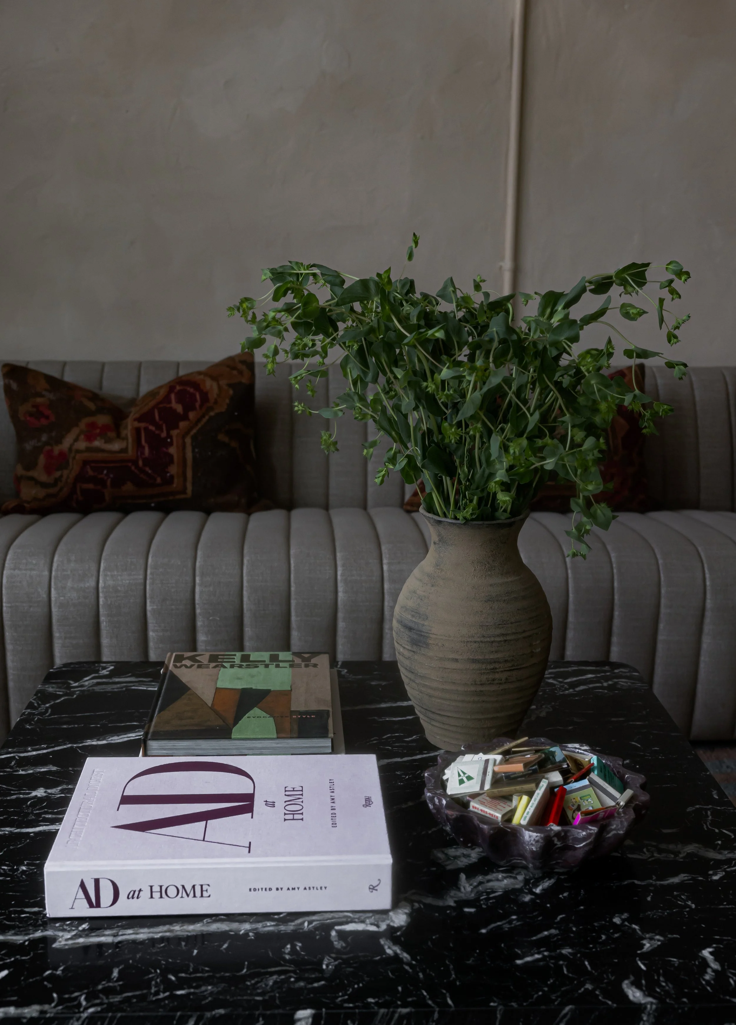 A marble-top table with a large book titled 'AD at Home' and a bowl of assorted candies, a potted green plant on a sofa with patterned pillows in the background.