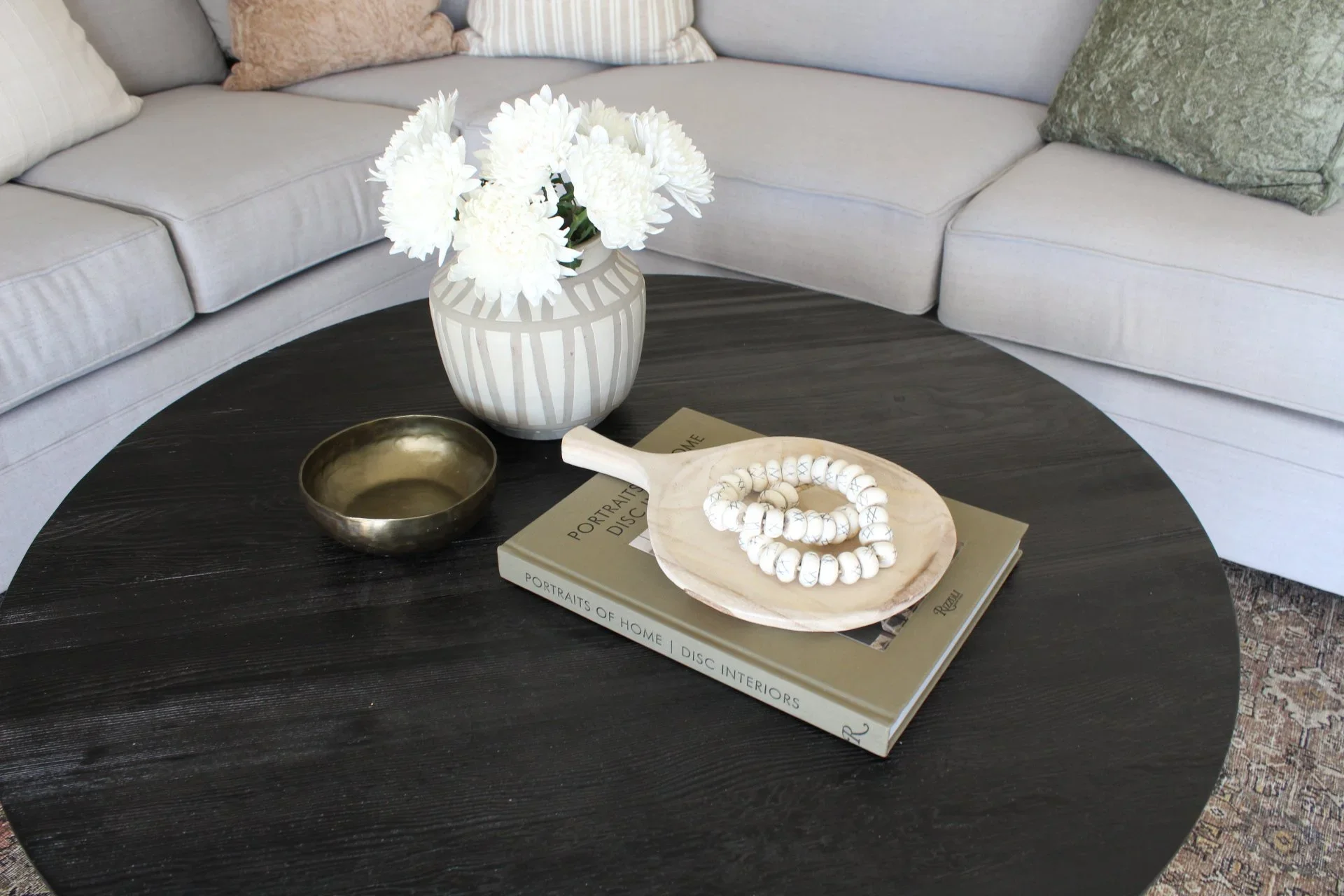A black round coffee table with a beige book titled 'Portraits of Home' on it, topped with white beaded jewelry on a shell-shaped dish, a small brass bowl, and a white vase with white flowers, set against a light-colored curved sofa with textured and