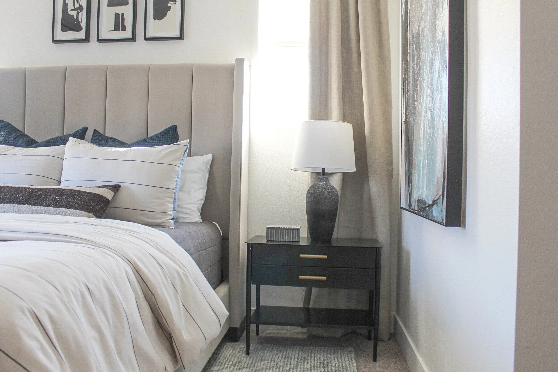 A bedroom with a bed, a black nightstand with a lamp and decorative box, wall art, curtains, and a window.
