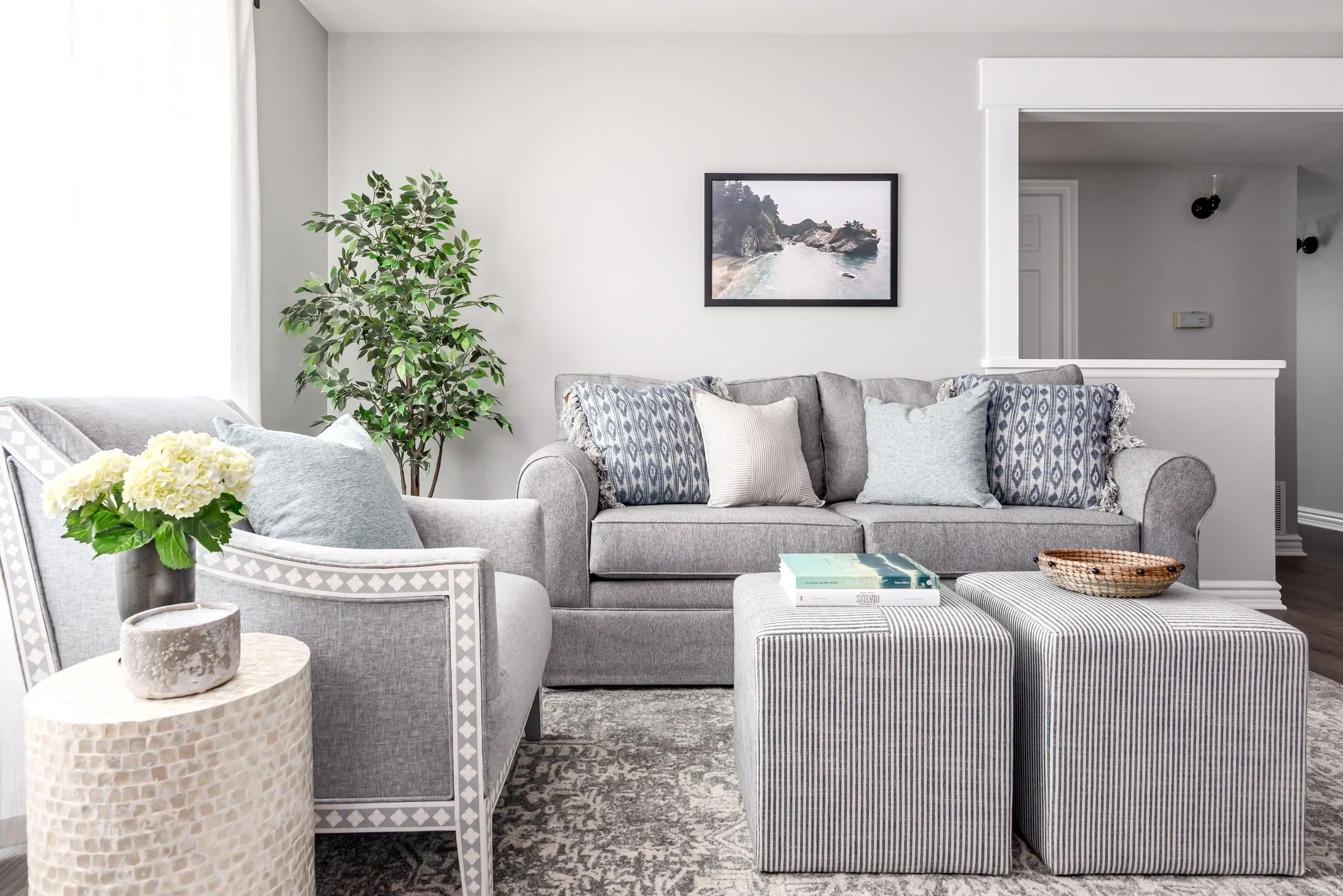 A cozy living room with light grey sofa, decorative pillows, striped ottoman, side table with vase of yellow flowers, large potted plant, wall art of a seascape, and a patterned area rug.