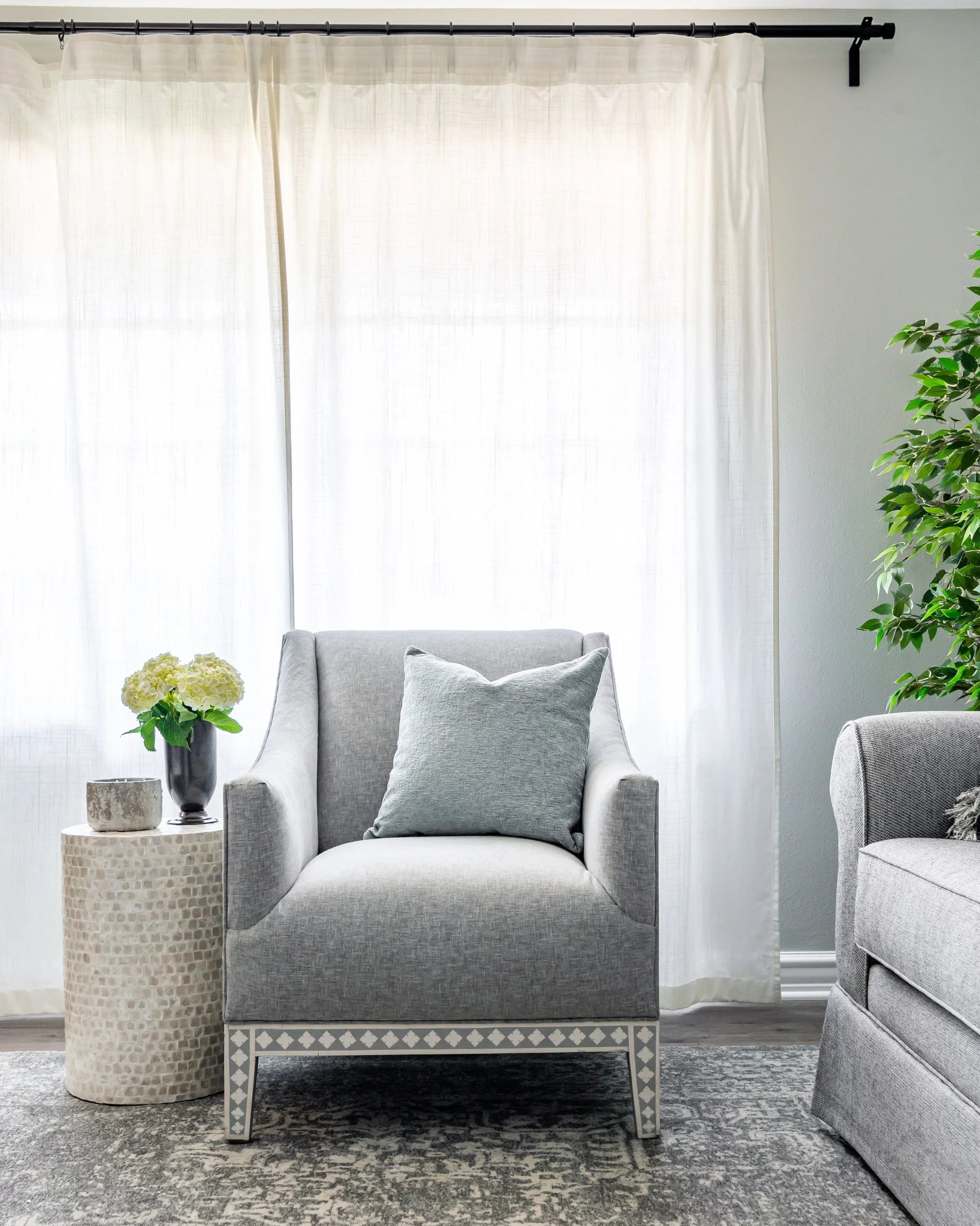 Living room with a gray armchair, a side table with a flower arrangement, white curtains, and a gray patterned rug.