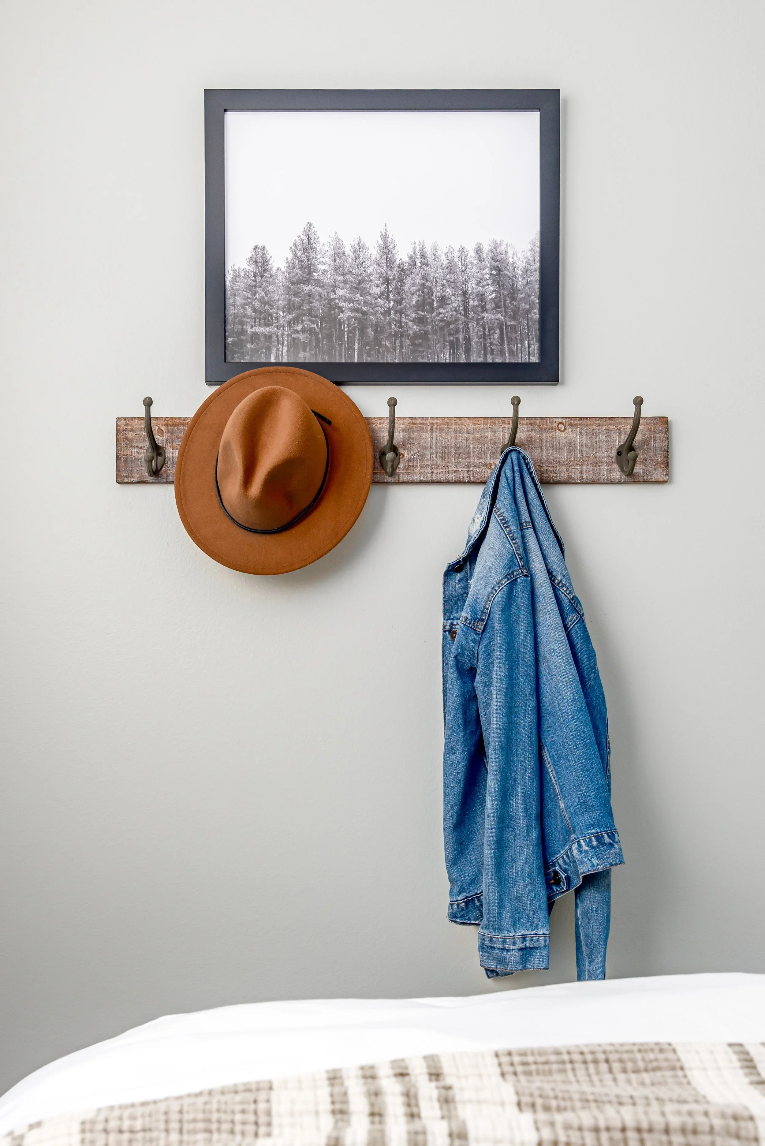 A brown wide-brimmed hat and a blue denim jacket hanging on hooks on a wooden board against a light-colored wall, with a black-and-white photograph of a snowy forest hanging above.
