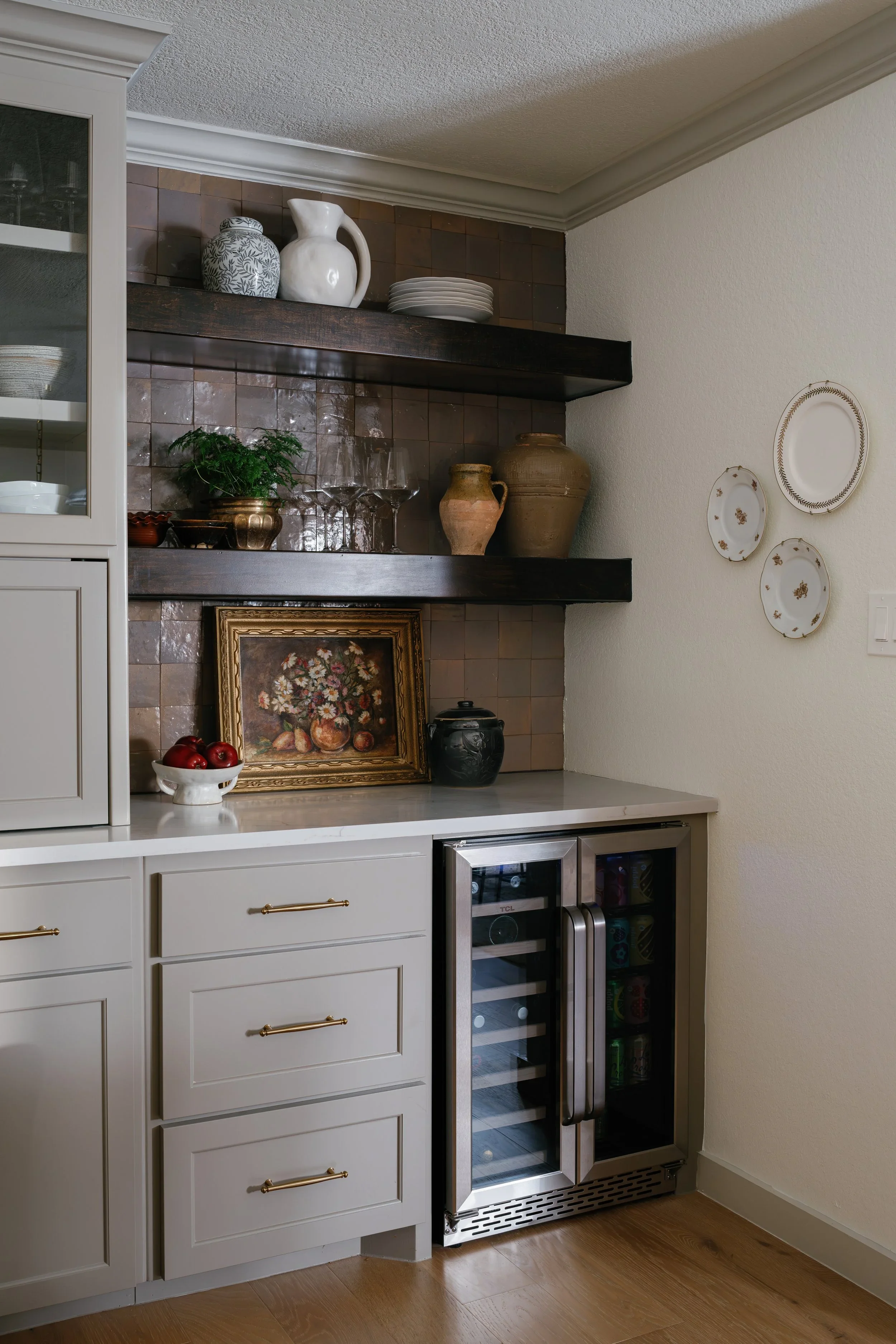 Section of a kitchen with white cabinetry, dark open shelves displaying vases, plates, and jars, a small wine cooler, and decorative wall plates.
