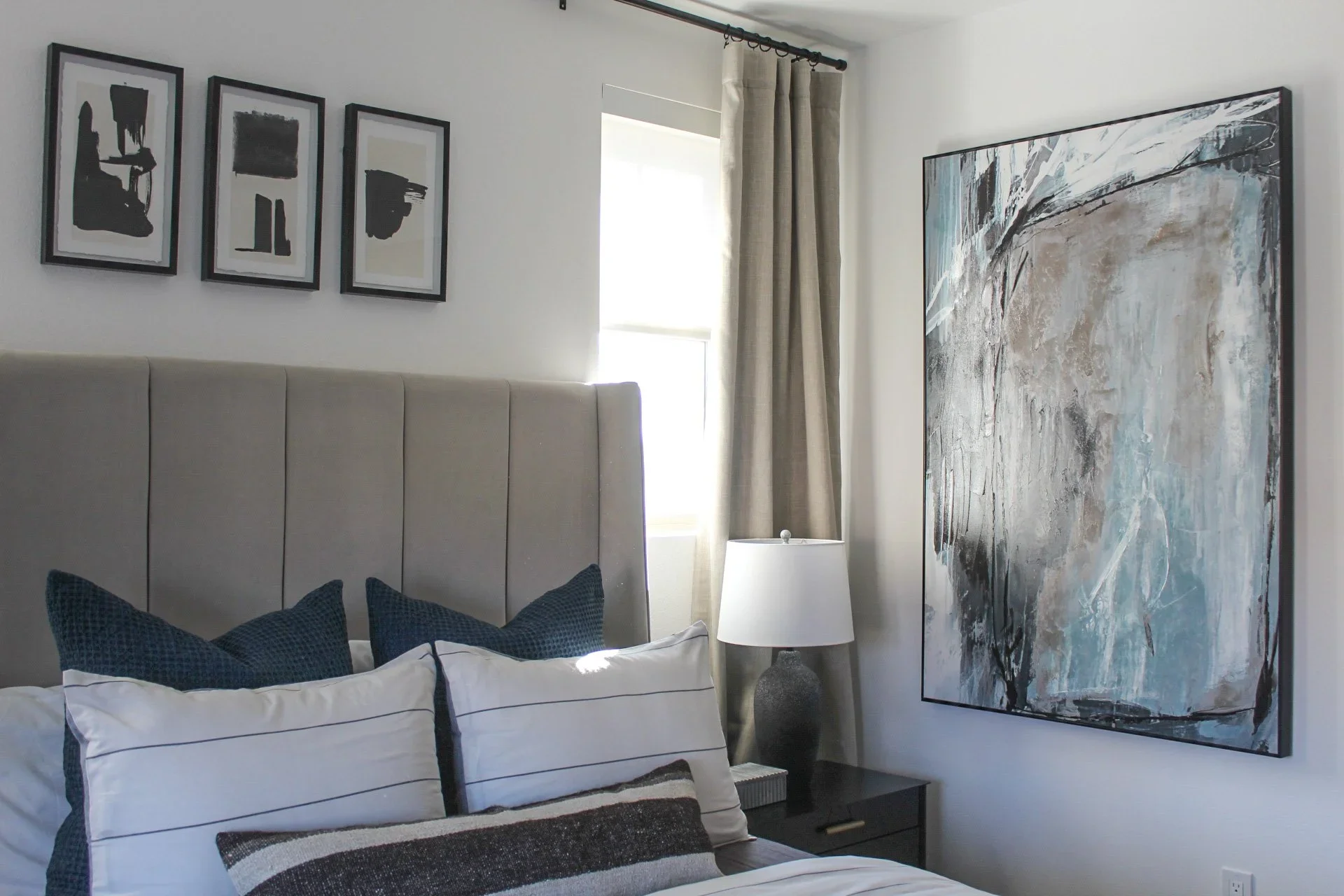 A bedroom with a beige upholstered headboard, white bedding with striped pillows, a bedside table with a table lamp, and wall art including three black abstract prints and a large textured painting. A window with beige curtains is visible, and sunlight streams into the room.