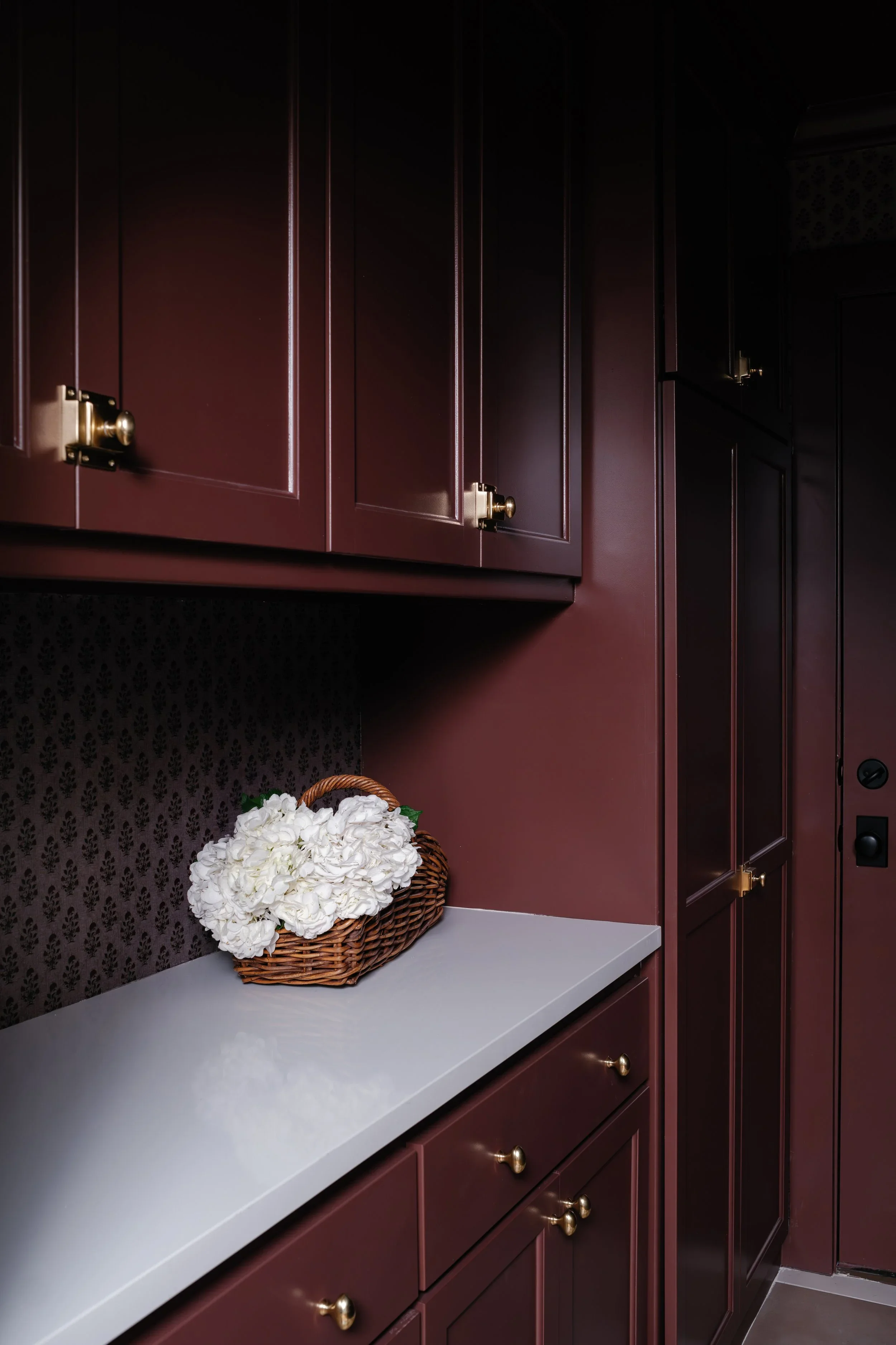 A kitchen cabinet with dark maroon doors and brass handles, a white countertop, and a basket of white hydrangea flowers on the counter.