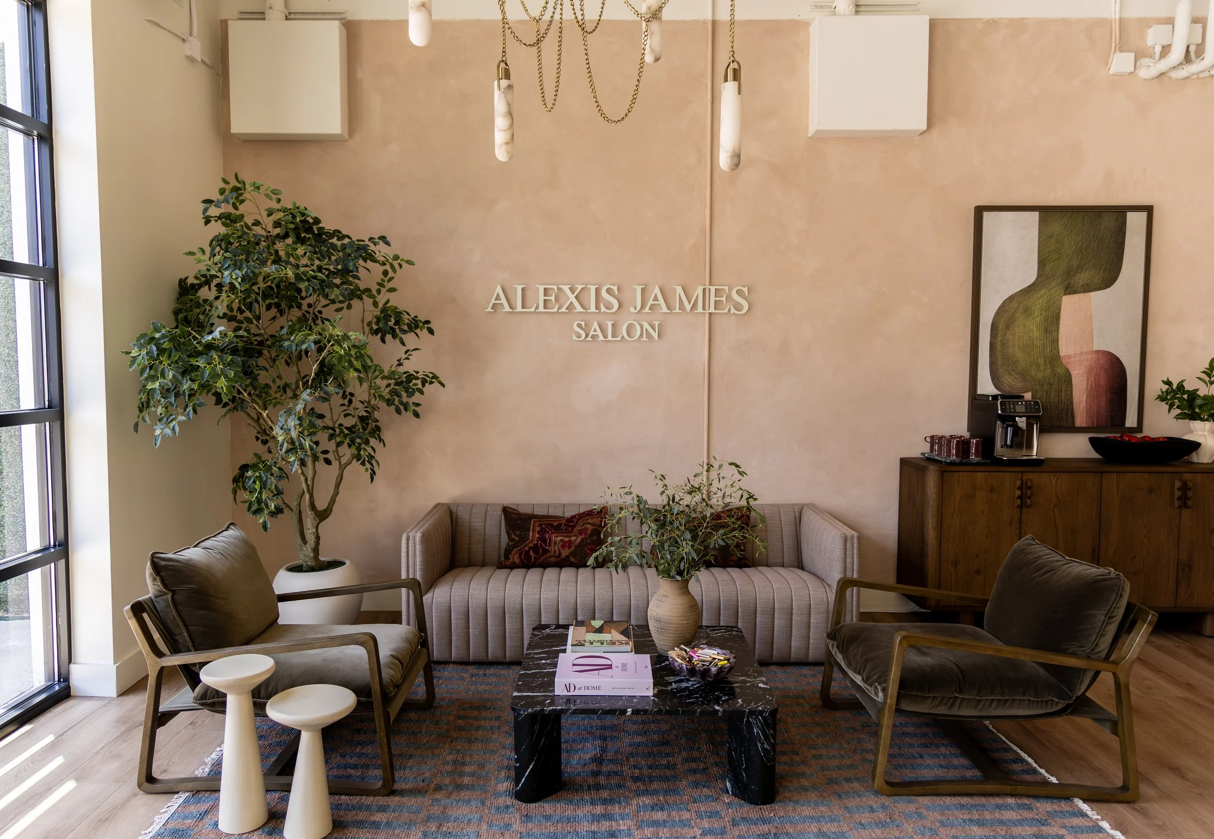 A salon waiting area with a beige sofa, two chairs, a black marble coffee table, and a large green potted plant. The wall behind has a sign that reads 'ALEXIS JAMES SALON' and decorative hanging lights. There is artwork on the wall and a window letti