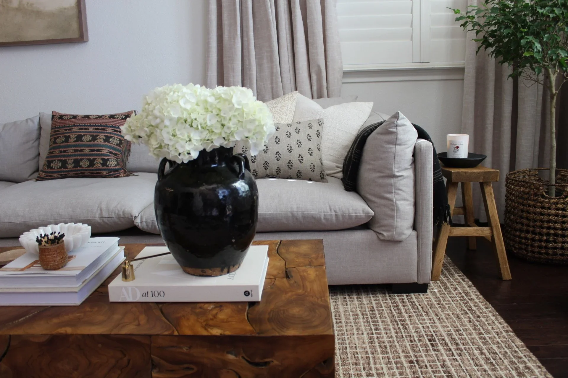 Living room with a beige sofa decorated with patterned cushions, a wooden coffee table with books and a black vase of white hydrangeas, a side table with a candle, curtains covering a window, a potted plant, and a woven basket.