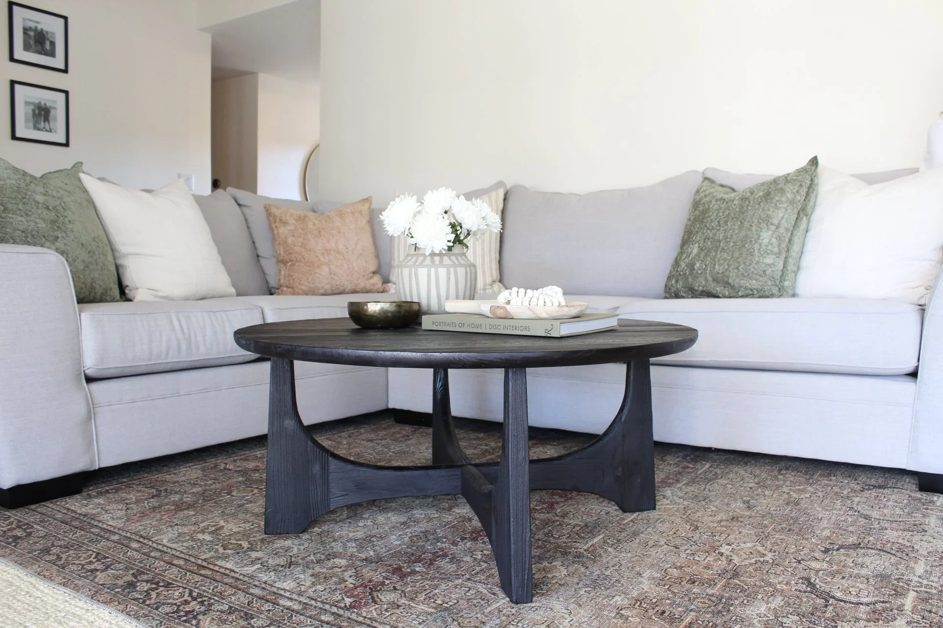 Living room with a white sofa, pillows, a dark wooden coffee table with a vase of white flowers, books, and decorative items. Framed black-and-white photographs on the wall and a patterned rug on the floor.