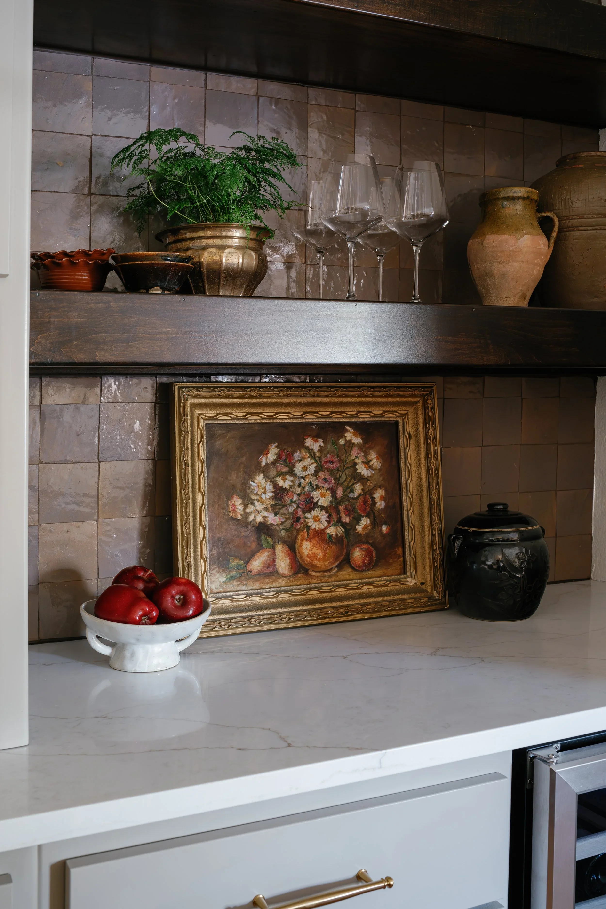 A kitchen countertop with a bowl of red apples, a framed still life painting of a vase with flowers, a black decorative jar, and a ceramic vase. Above the countertop is a wooden shelf holding glassware, plants, and pottery, with beige and brown tiled