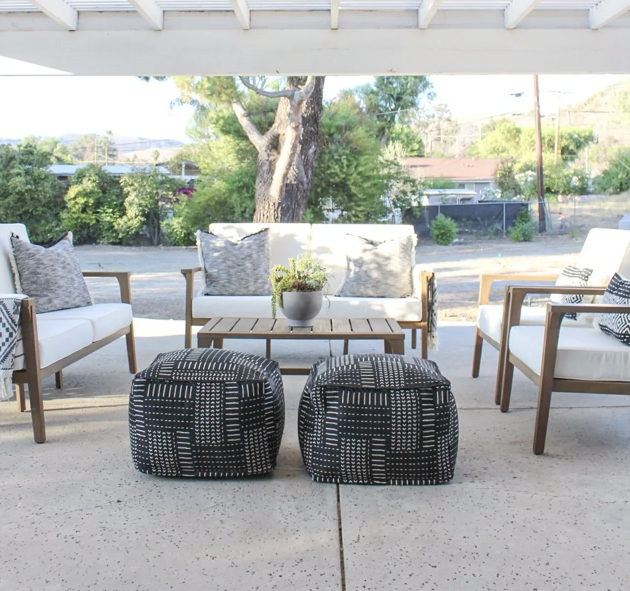 Outdoor patio furniture with white cushioned chairs, a white cushioned loveseat, and two black and white patterned ottomans around a wooden coffee table with a potted plant, set against a backdrop of trees and a backyard.