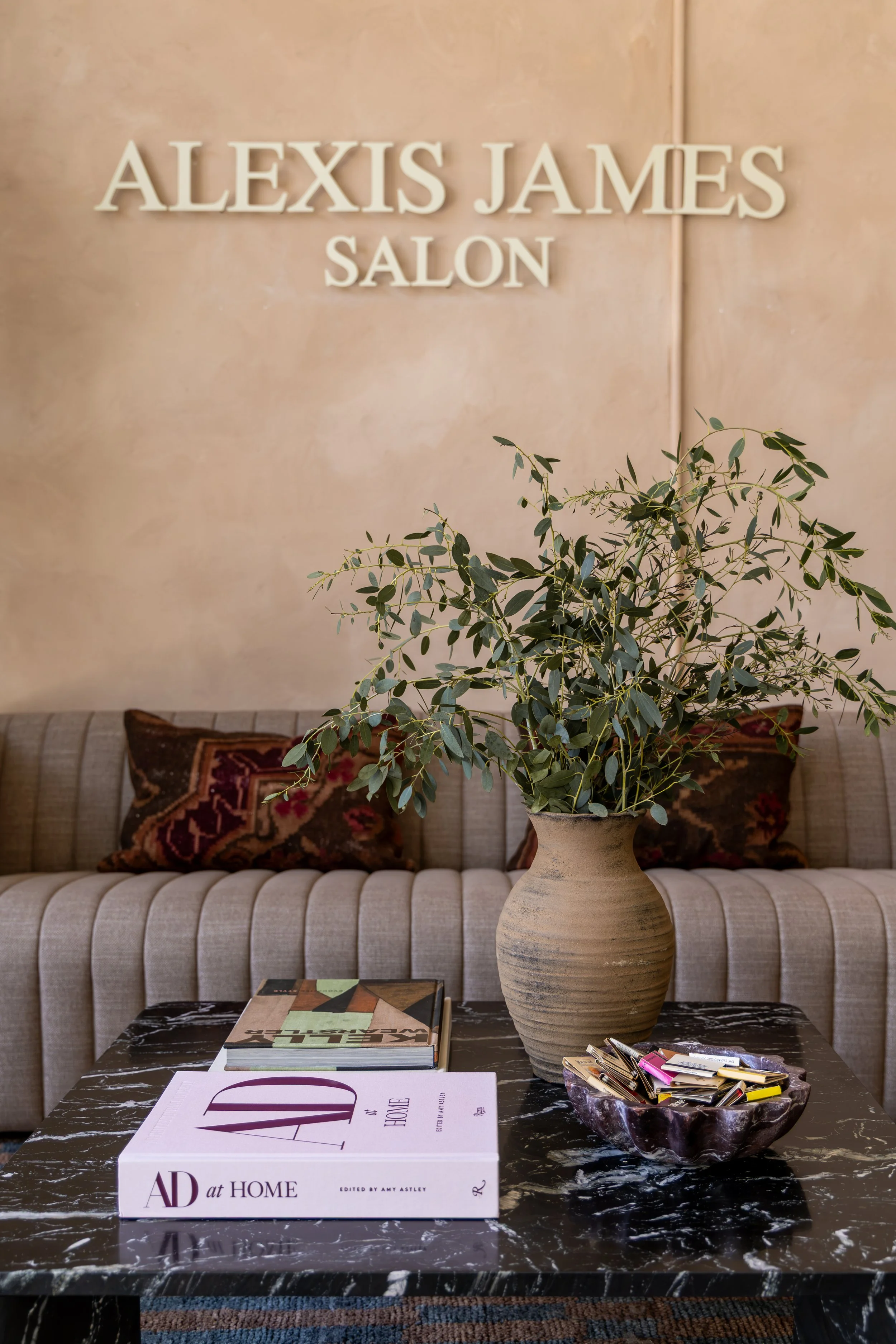A coffee table in a living room with a large decorative vase containing green leafy branches, a stack of magazines, a purple and white book titled "AD at HOME," and a marble-like tray holding assorted clips and office supplies. In the background, a b