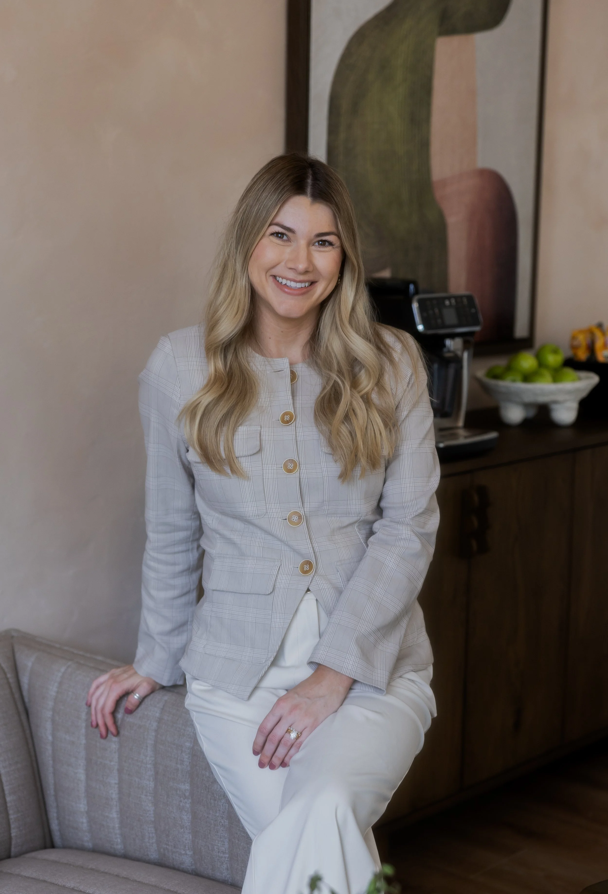 A smiling woman with long wavy blonde hair wearing a beige plaid blazer and white pants, sitting on a beige armchair in a room with a modern abstract painting and a bowl of green apples in the background.