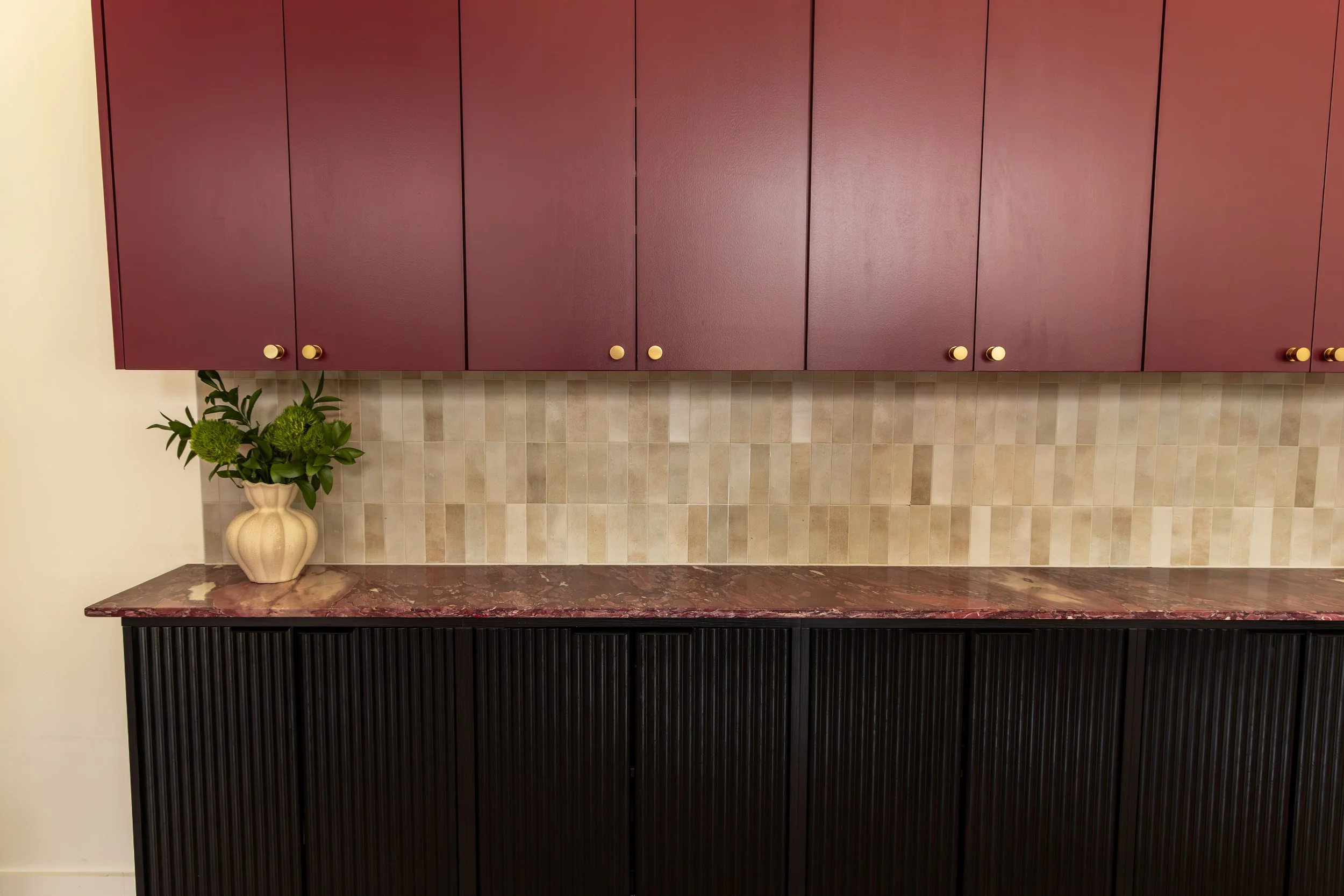 A kitchen with mauve-colored cabinets, a marble countertop, beige tile backsplash, a black textured cabinet below, and a potted plant in a cream-colored vase.