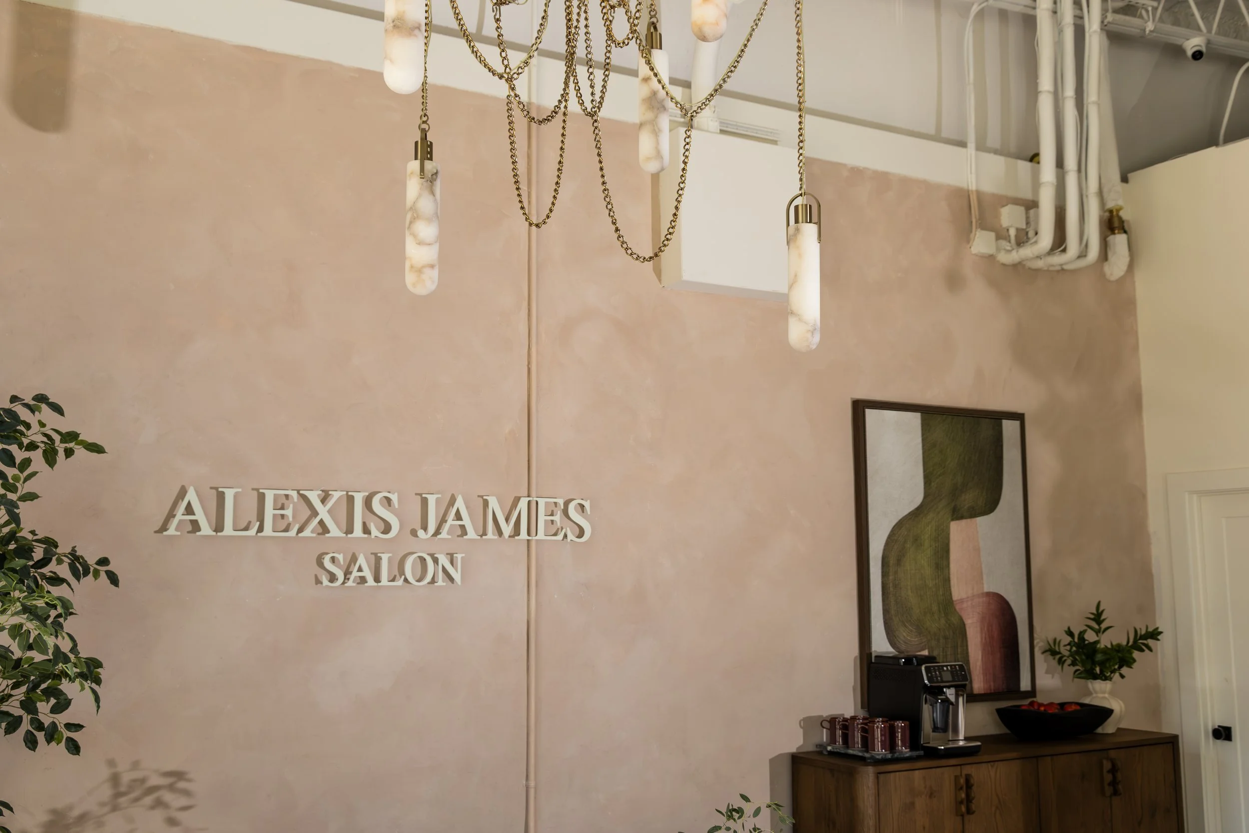 Interior of a salon with a pink wall, a wooden reception desk, framed mirror, and hanging chains with marble-like handles.