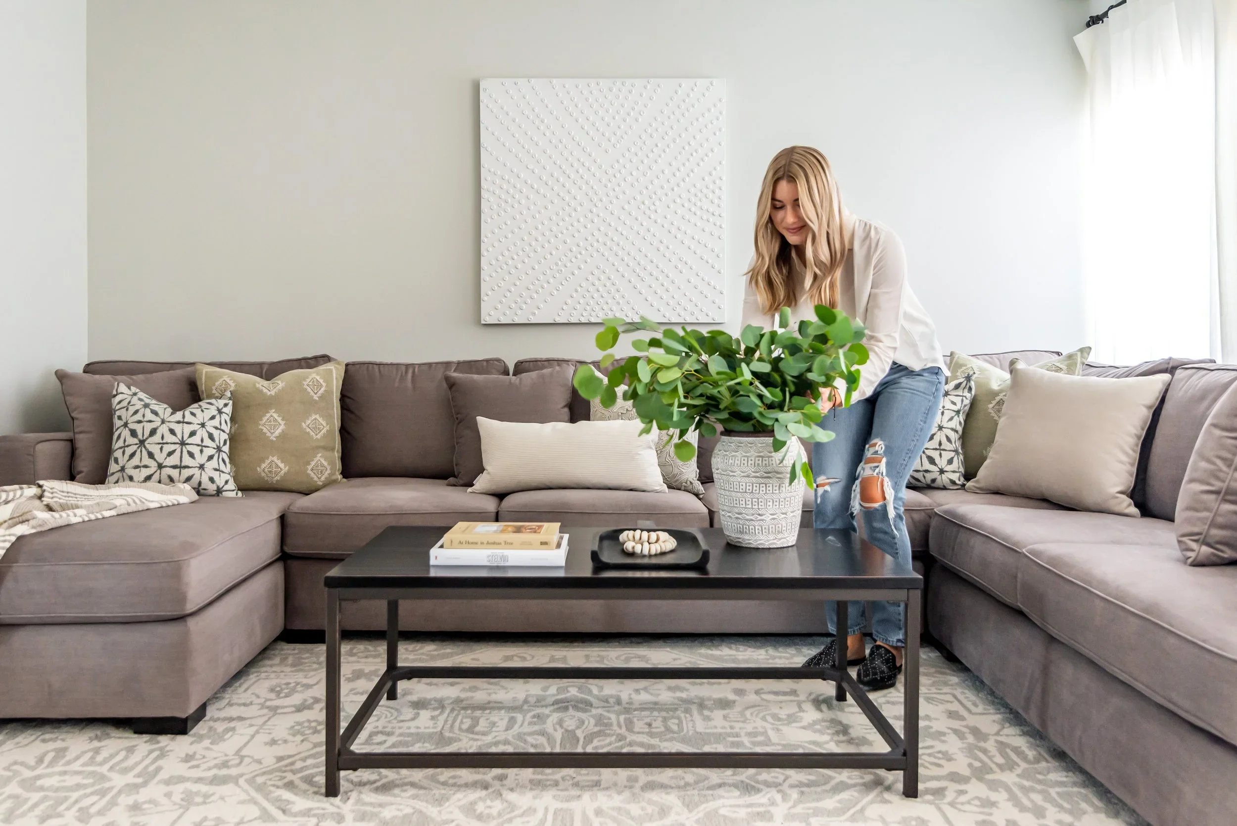 A woman with long blonde hair, wearing a white shirt and ripped jeans, is arranging a green potted plant on a black coffee table in front of a gray sectional sofa with patterned and solid-colored pillows. There is a white textured artwork on the ligh