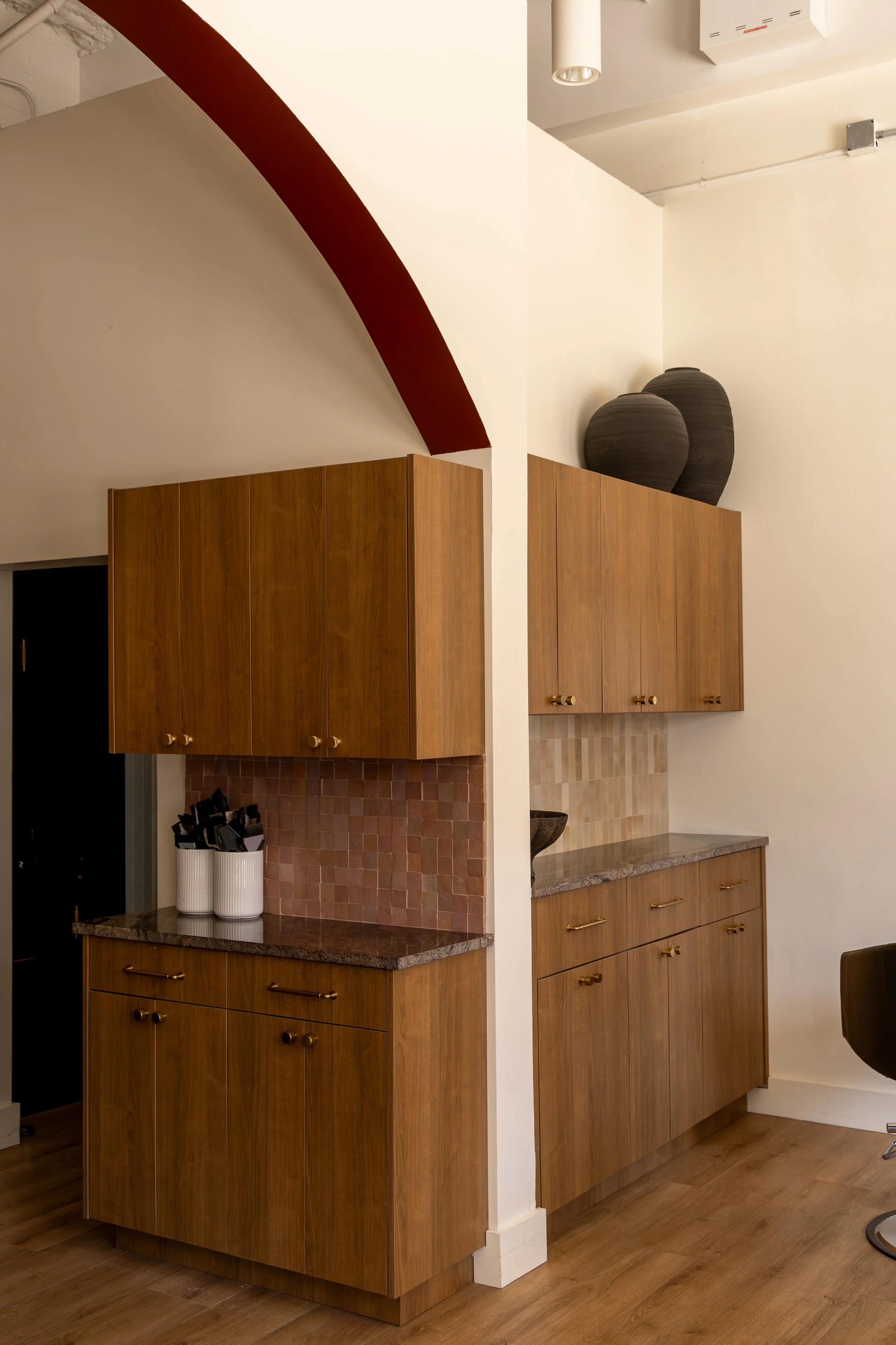 A kitchen with wooden cabinets, a granite countertop, and decorative vases on top. There are hanging copper utensils and a tiled backsplash.