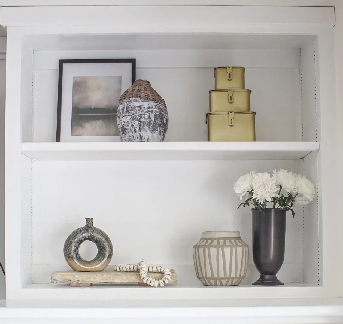 White display shelf with decorative objects including a framed picture, a woven vase, three yellow tin boxes, a round metallic vase, a beaded necklace, a patterned vase, and a black vase with white flowers.