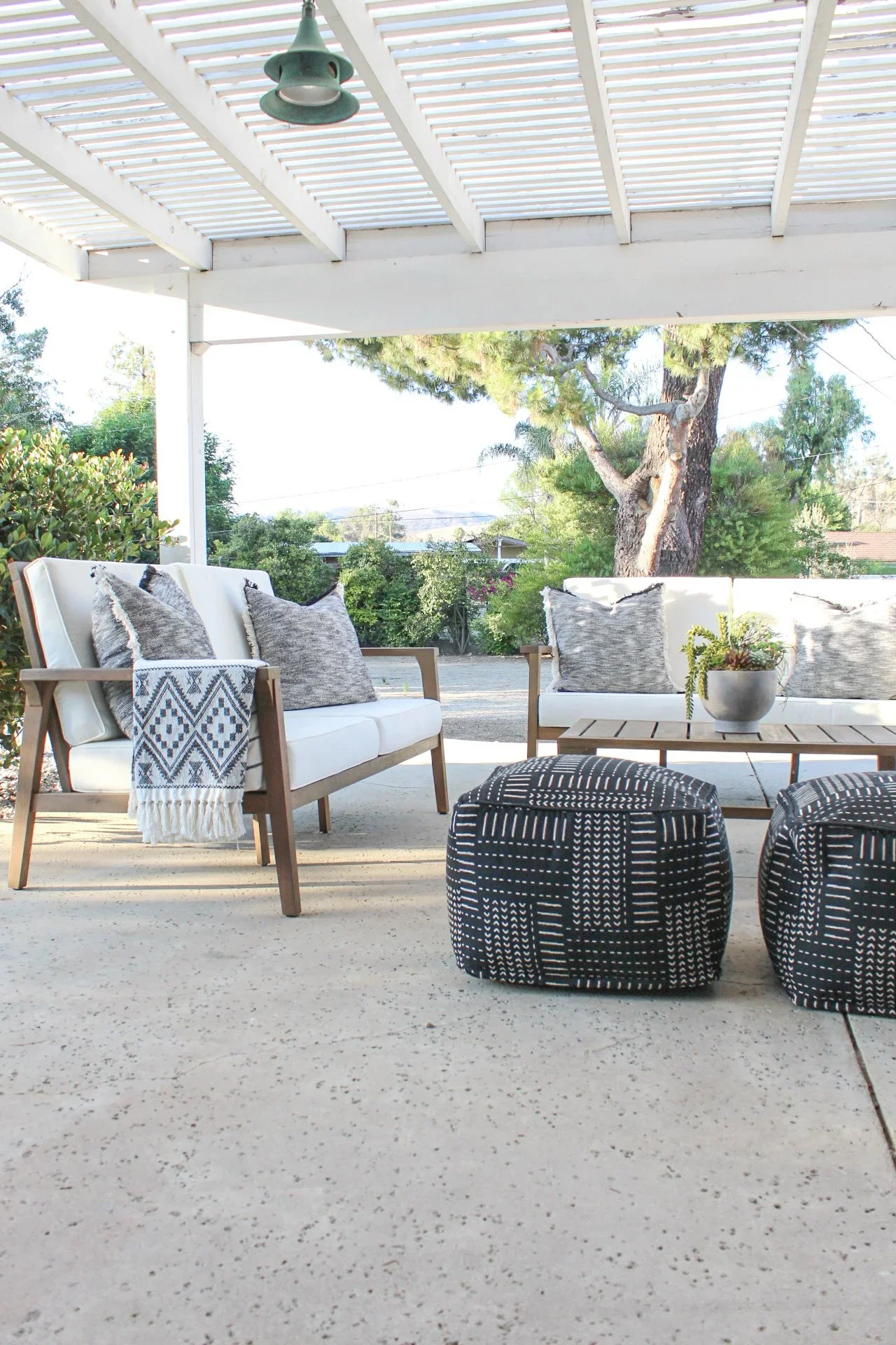Outdoor patio with white seating, patterned throw pillows, a small coffee table with a potted plant, and two black-and-white patterned ottomans. A large tree and greenery are visible in the background.