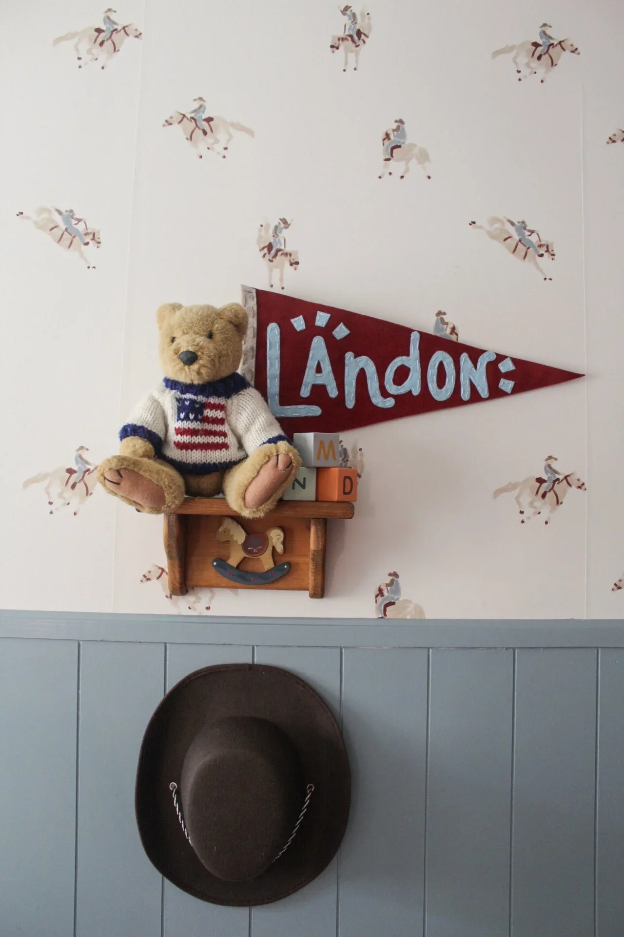 A wall with a western-themed wallpaper featuring cowboy riders on horses. There is a red pennant with blue letters spelling 'L'Àndon', a teddy bear wearing a knitted sweater, wooden blocks, a small wooden shelf, and a brown cowboy hat hanging below, all on a tiled wall.