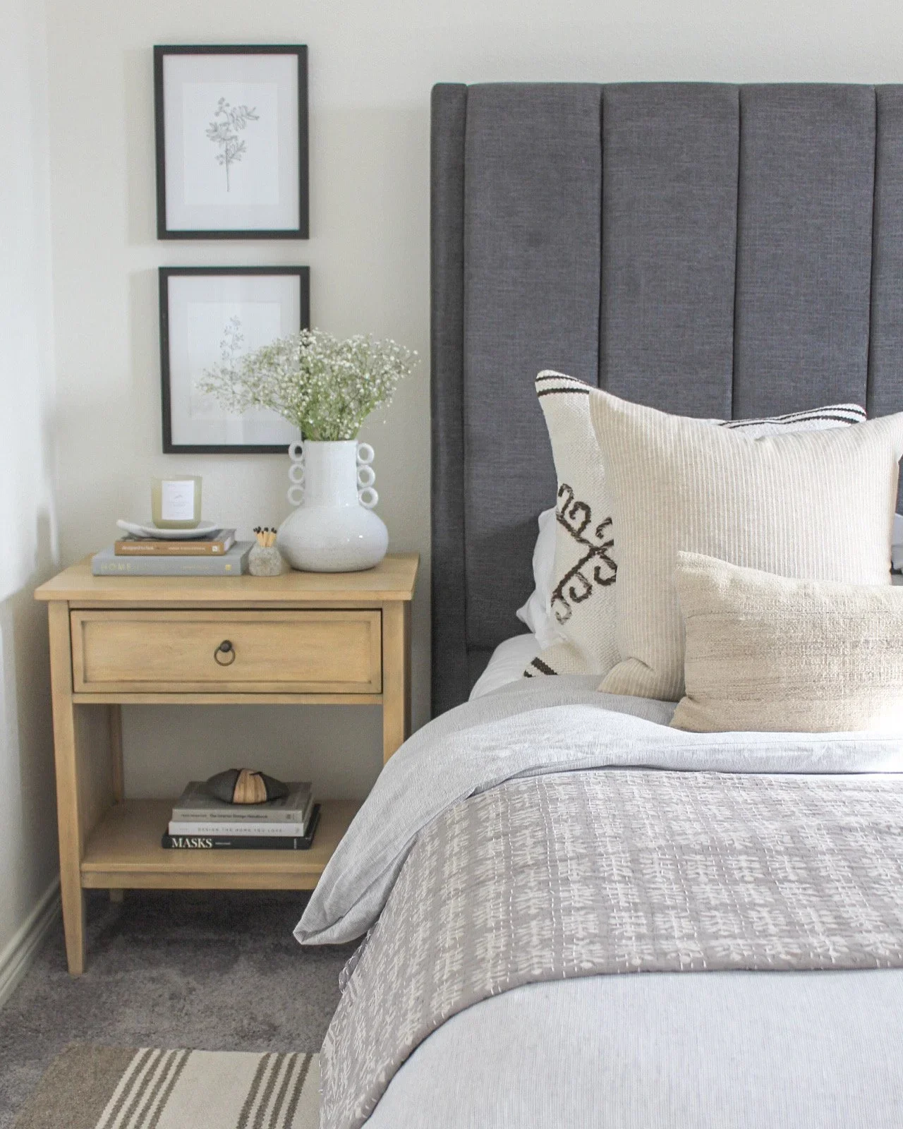 A bedroom with a grey upholstered headboard, beige pillows, a nightstand with a vase of white flowers, books, and decor, and framed black-and-white botanical art on the wall.