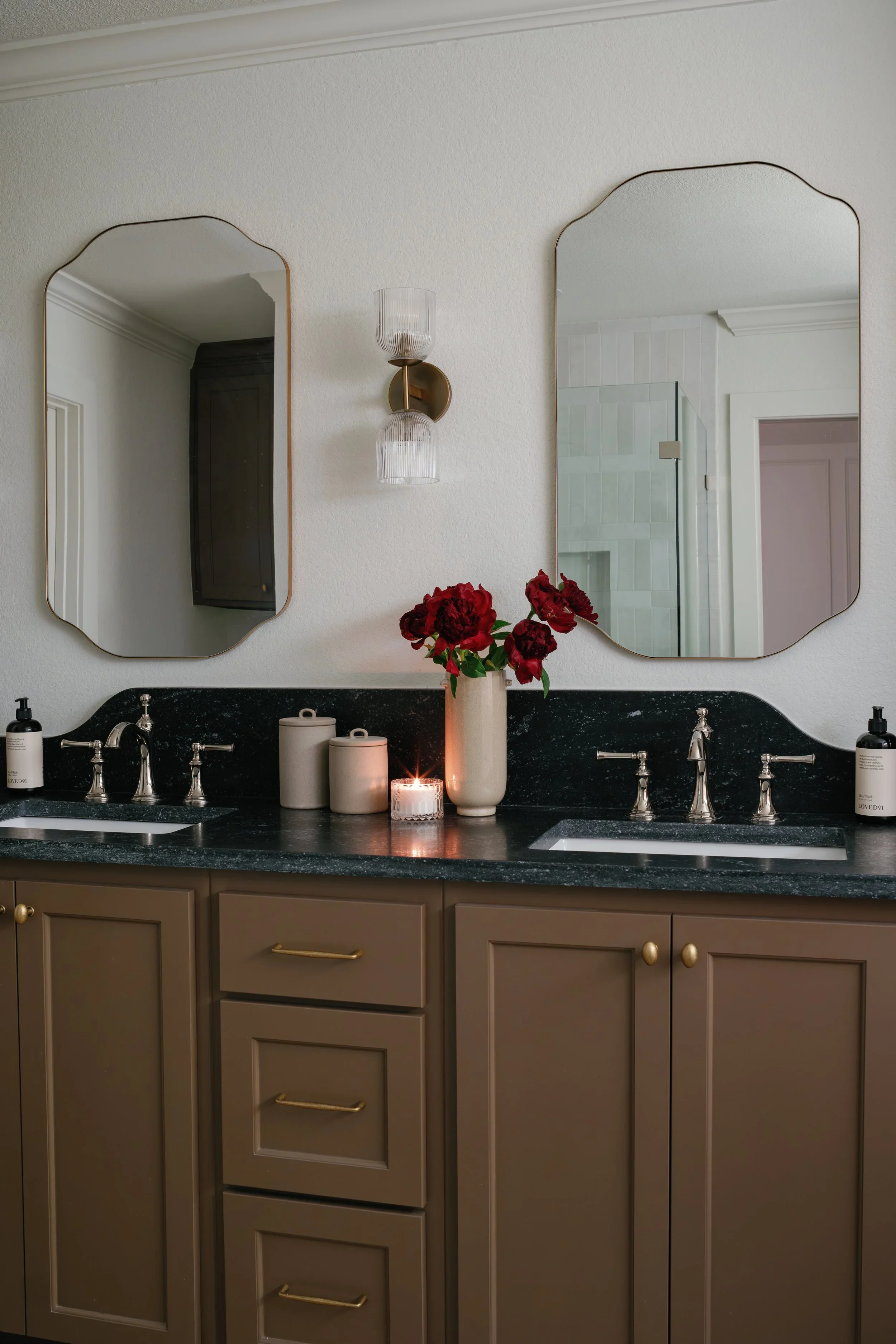 Bathroom vanity with two mirrors, beige cabinets with gold handles, black countertop, two sinks, soap dispensers, lit candle, vase with red flowers, wall sconce, and a glimpse of a shower and door in the background.