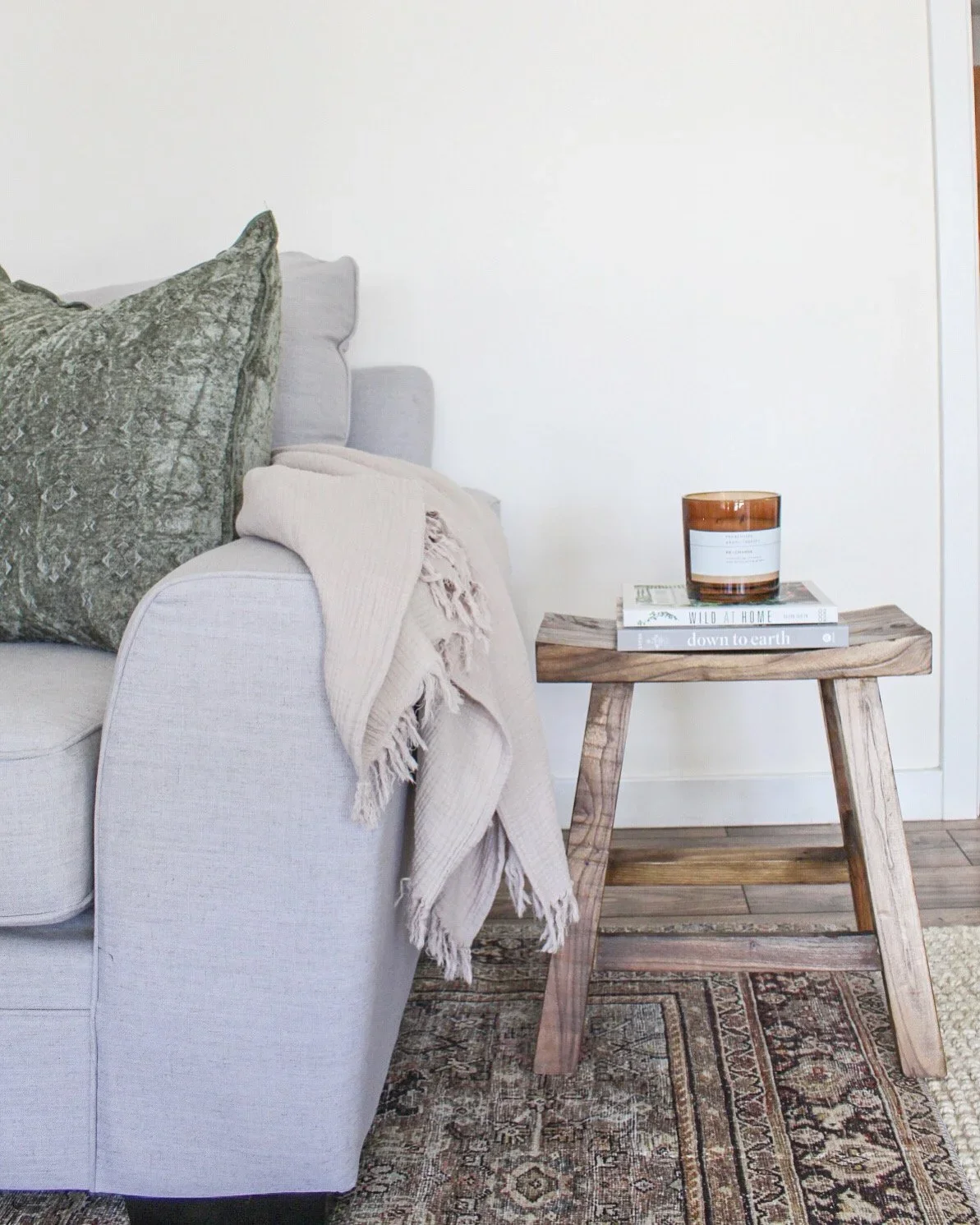 A cozy living room corner featuring a light-colored sofa with textured pillows and a blanket, next to a rustic wooden side table holding a candle and books, with a patterned rug on the wooden floor.