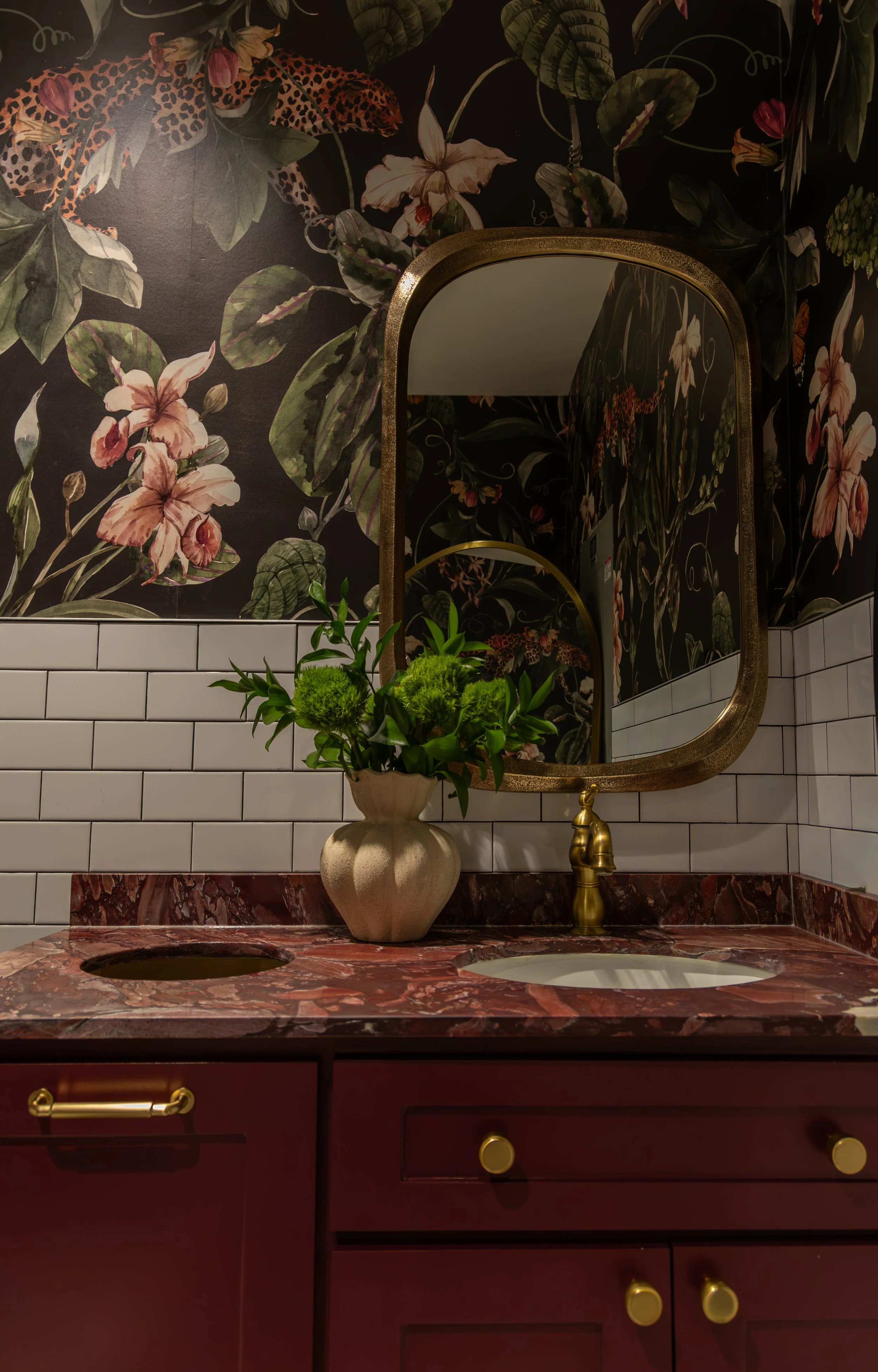 Bathroom vanity with a red marble countertop, a gold faucet, a white vase with green flowers, a large arched mirror, a floral wallpaper with leopards, flowers, and leaves, and white subway tiles on the wall.