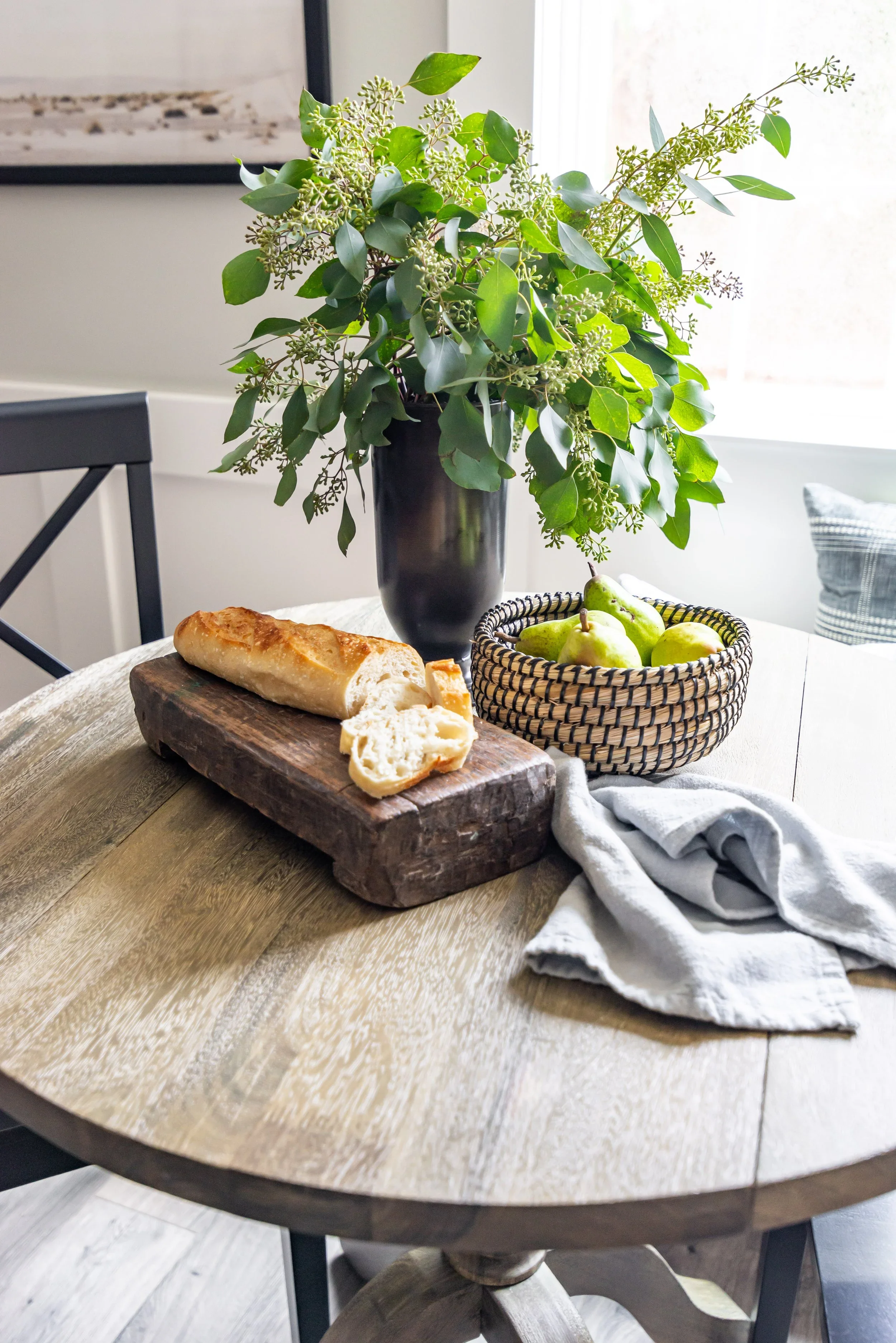 A round wooden dining table with a loaf of bread on a wooden cutting board, a basket of green pears, and a dark vase with green leafy branches, near a window in a bright room.
