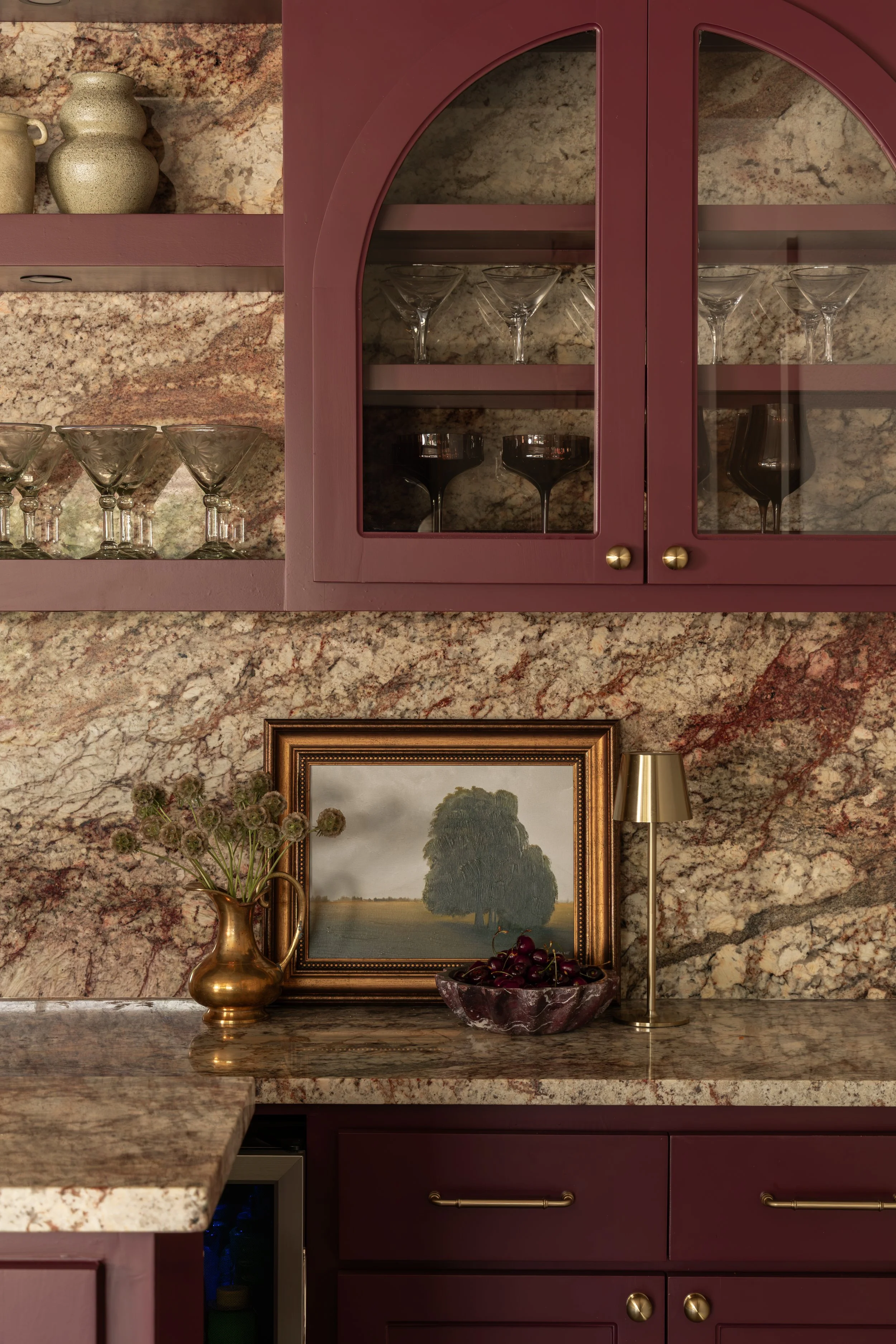 A kitchen with burgundy cabinets, granite countertops, and a marble backsplash. Open glass-front cabinet displaying glassware, and decorative items such as a vase with flowers, a framed painting of a tree, a bowl of cherries, and a small lamp.