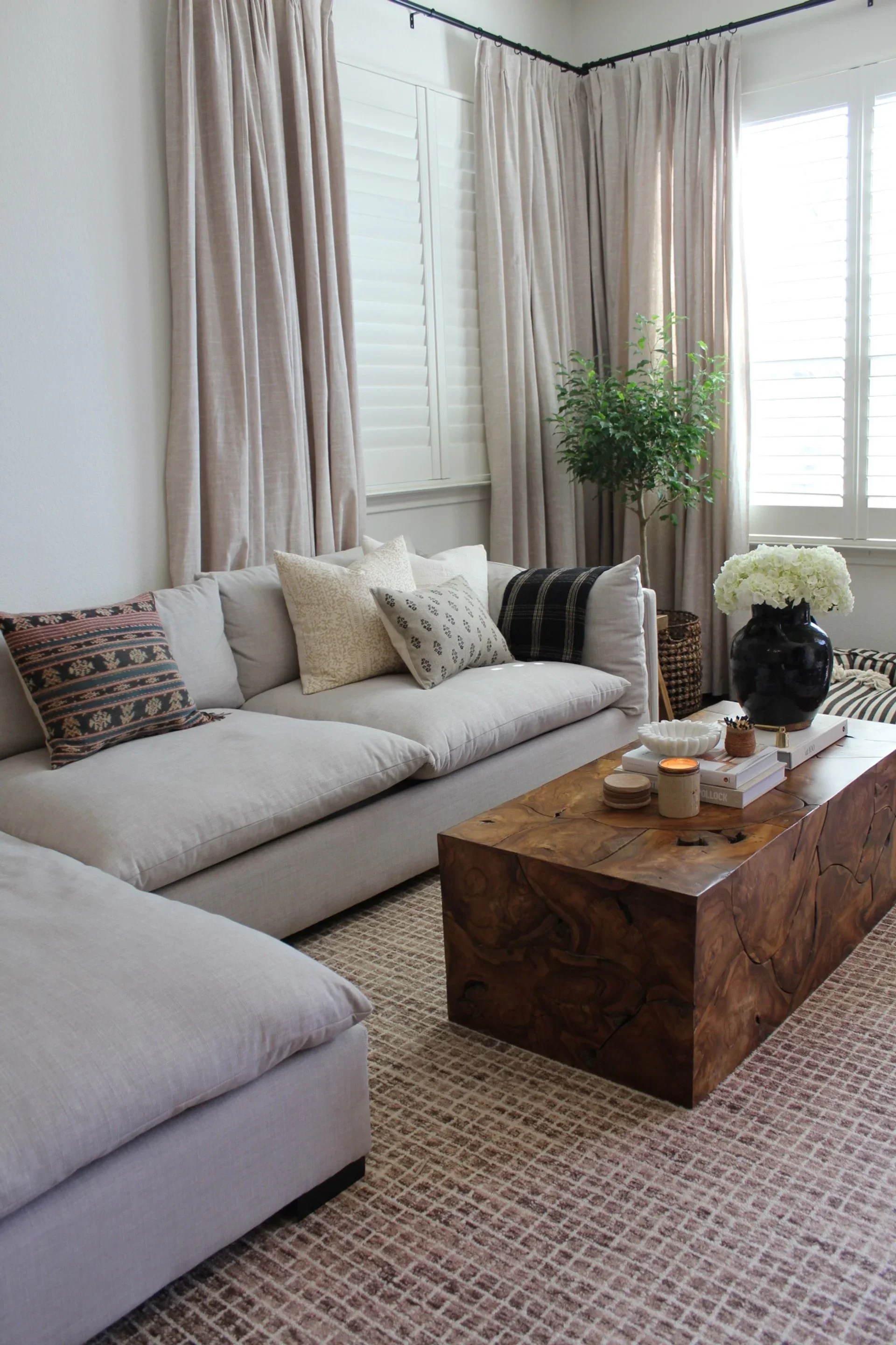 Living room with beige sofas, decorative pillows, a wooden coffee table, black vase with white flowers, and large windows with beige curtains.
