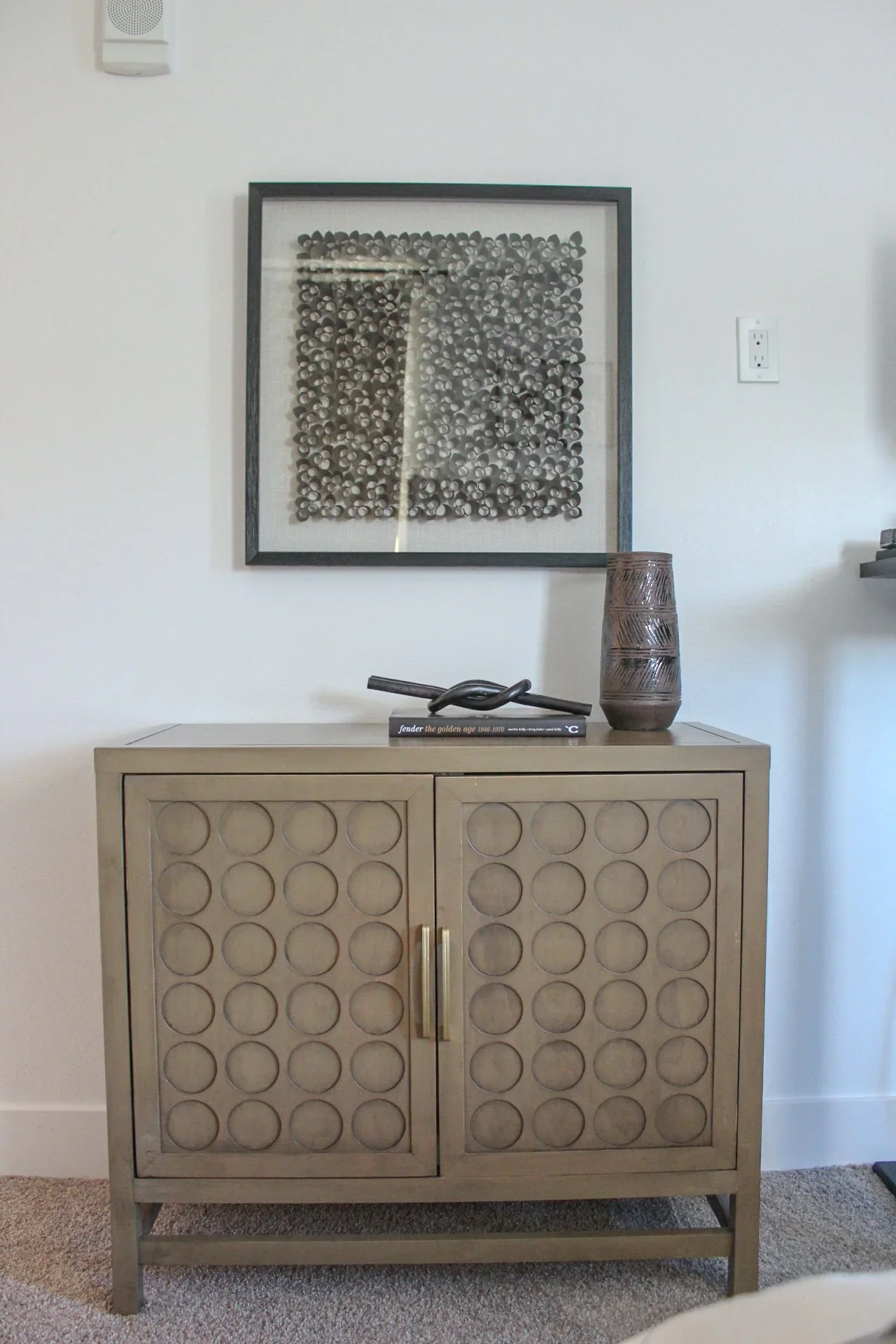 A beige cabinet with circular pattern on the doors, decorated with a black sculpture, a tall decorative vase, and a book titled 'Fender the golden age 1946-1970' on top, against a white wall with a framed collage of small black and white circular obj