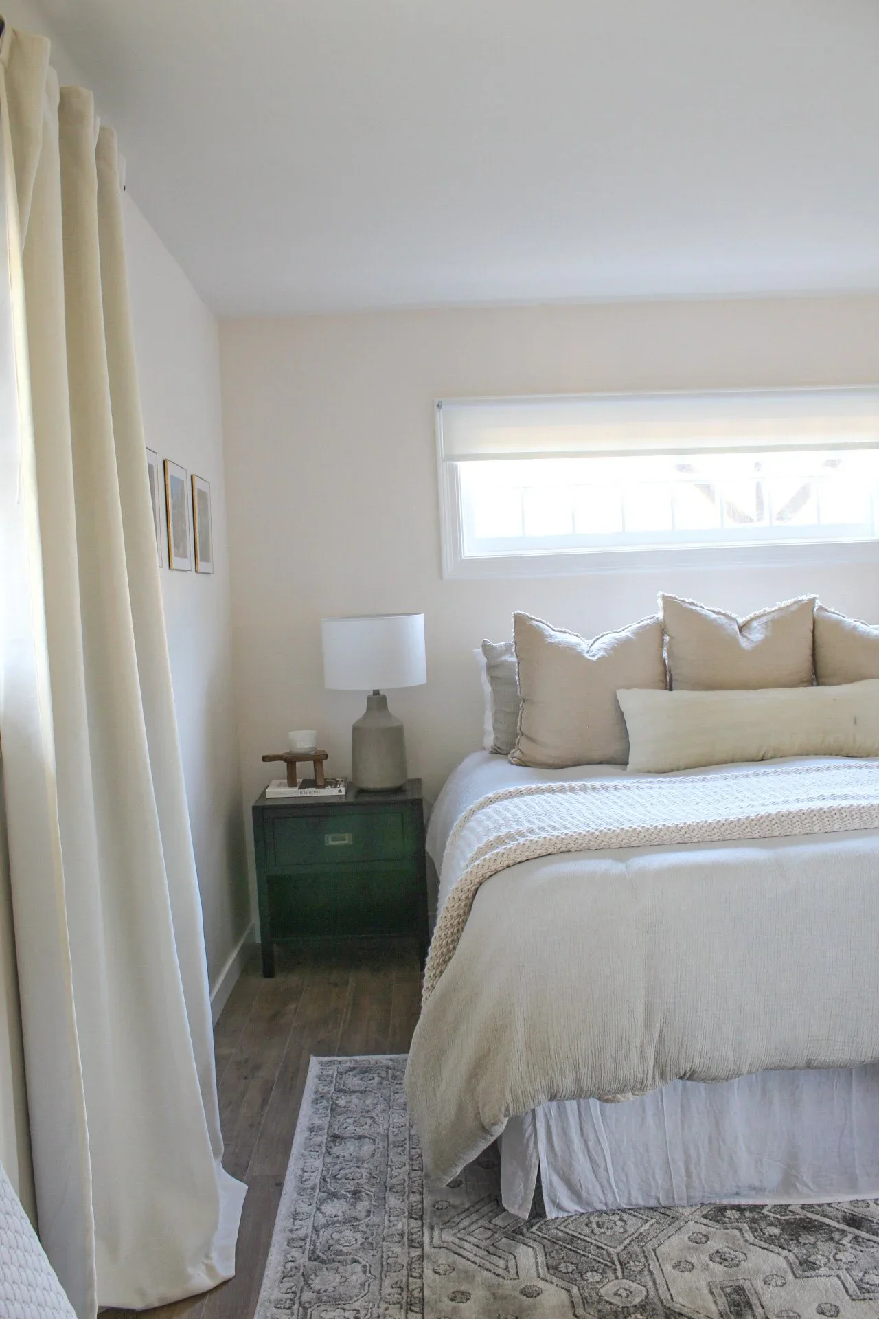 A bedroom with a bed, beige pillows, a nightstand with a lamp, a window with blinds, and cream-colored curtains.