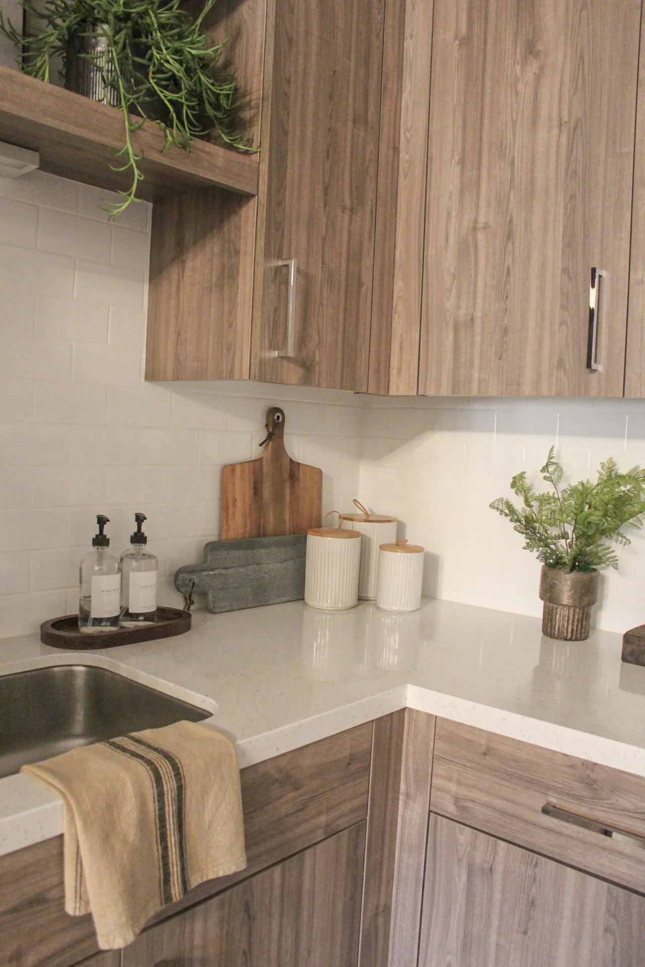 Kitchen with wooden cabinets, white countertop, potted plants, soap dispensers, and kitchen towels.