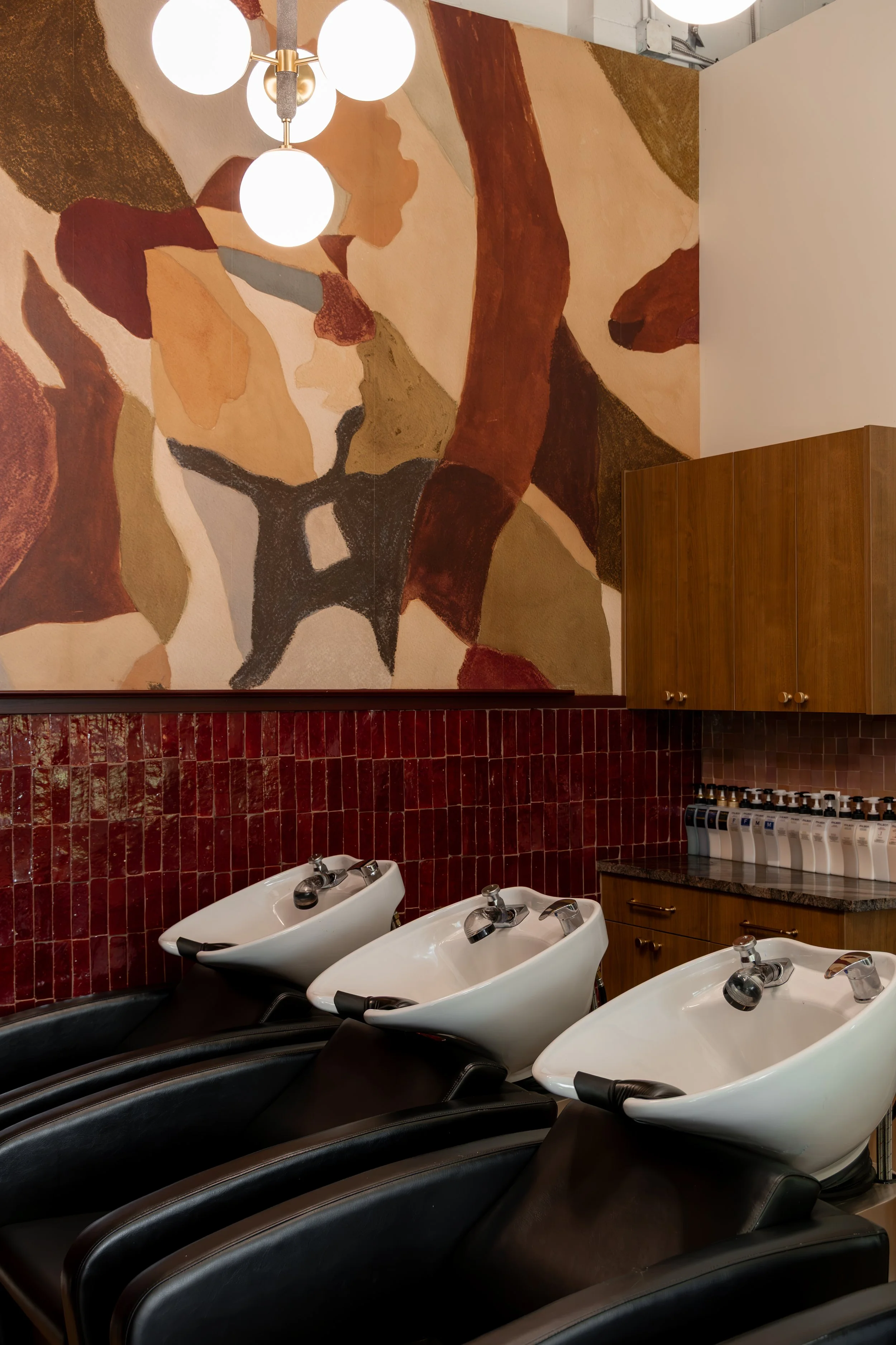 A row of three white shampoo sinks in a hair salon with black chairs in front of them, a red brick wall, a colorful abstract mural, and a wooden cabinet with small bottles on a countertop.